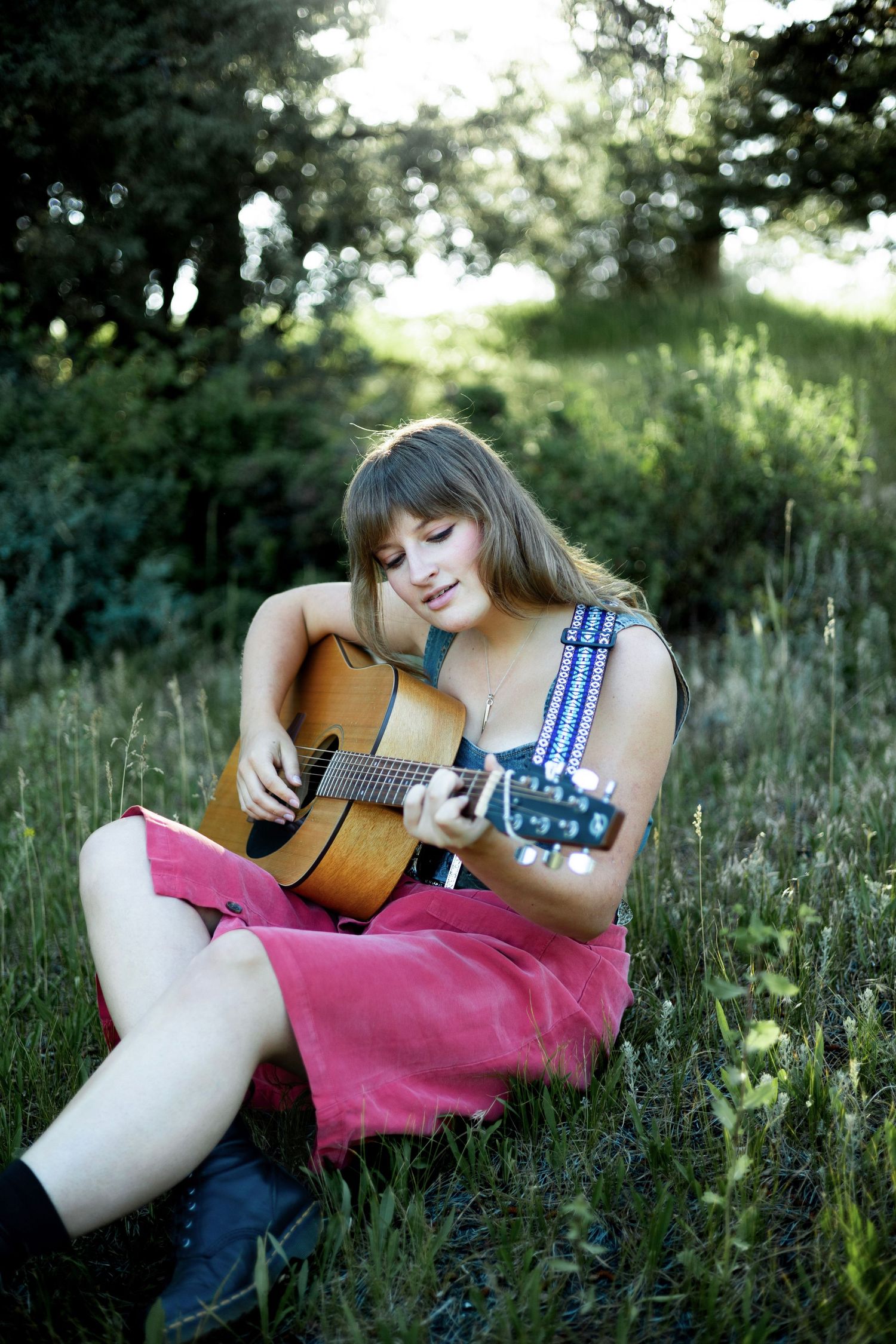 Young woman in pink dress playing acoustic guitar while sitting on grass in a lush green outdoor setting.