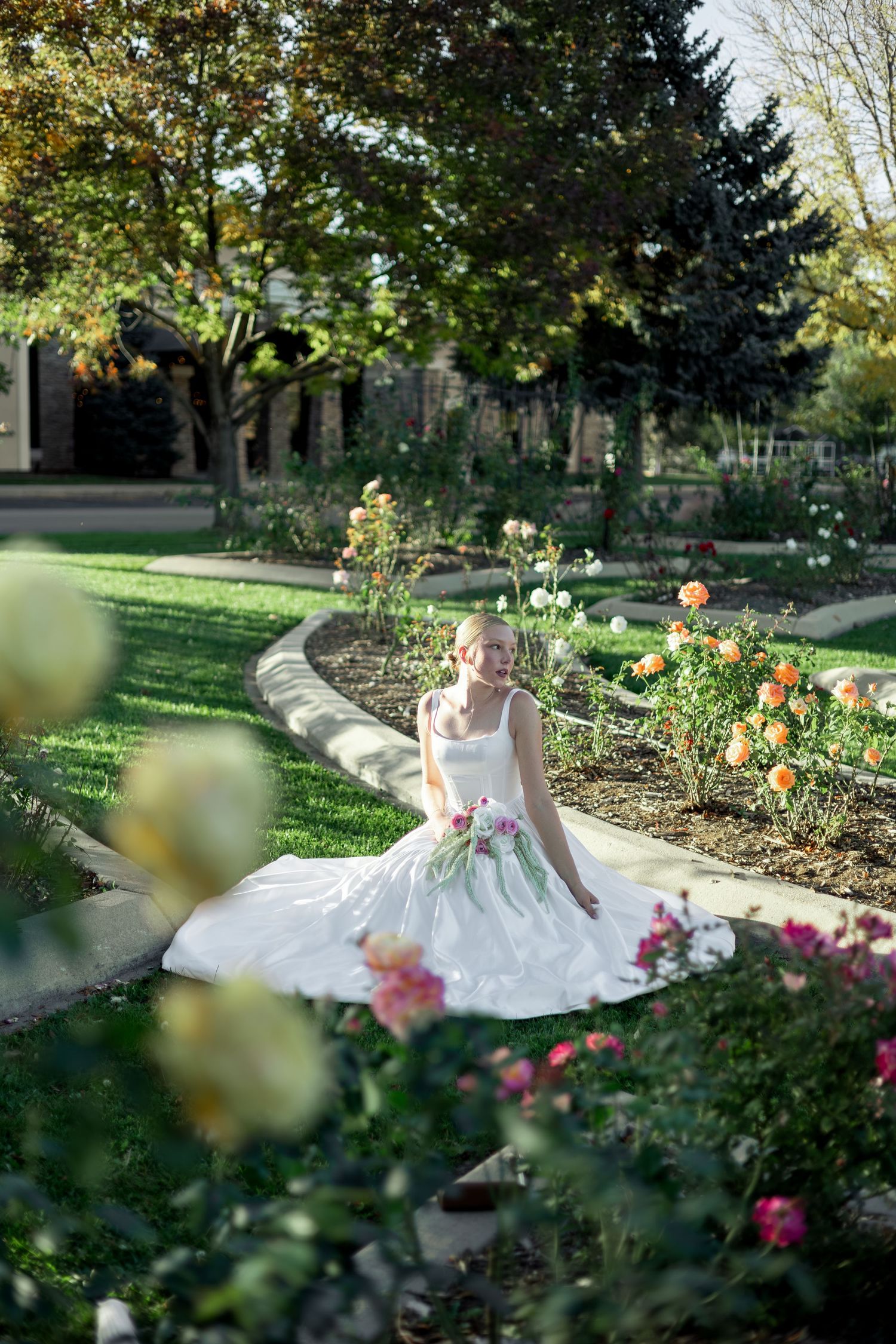 Bride in white gown with floral accents poses in a lush rose garden on a sunny day.