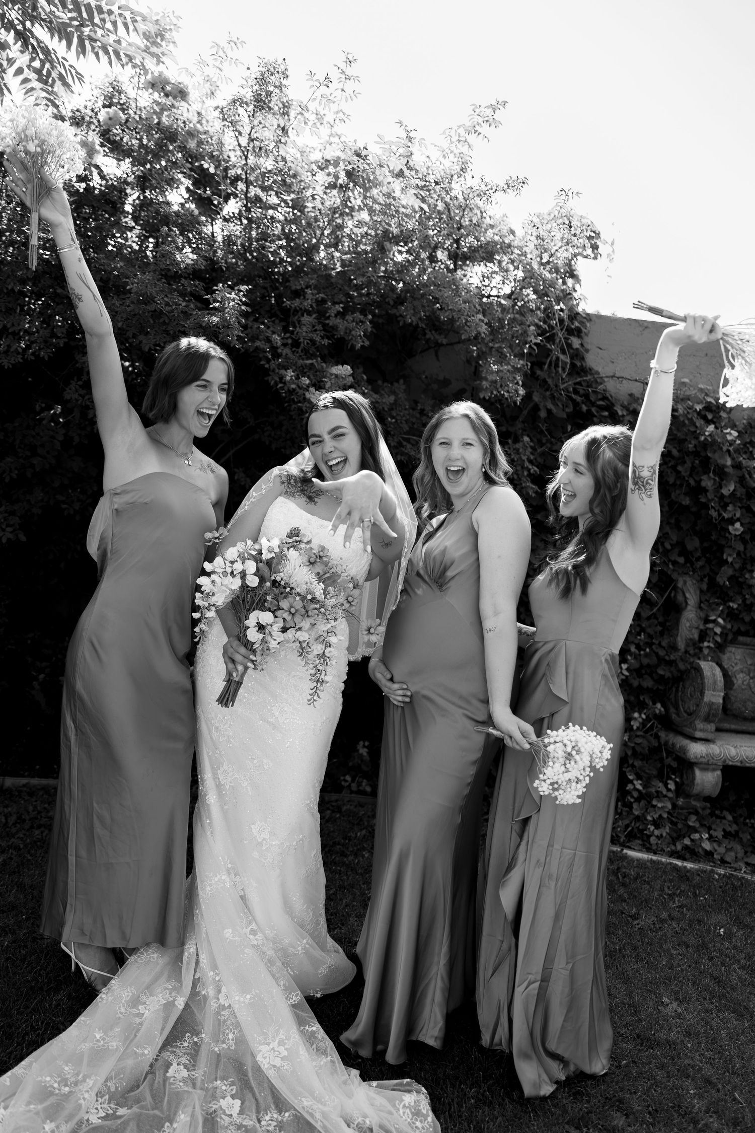 Bride and three bridesmaids in sage gowns celebrate joyfully outdoors in black and white photo.
