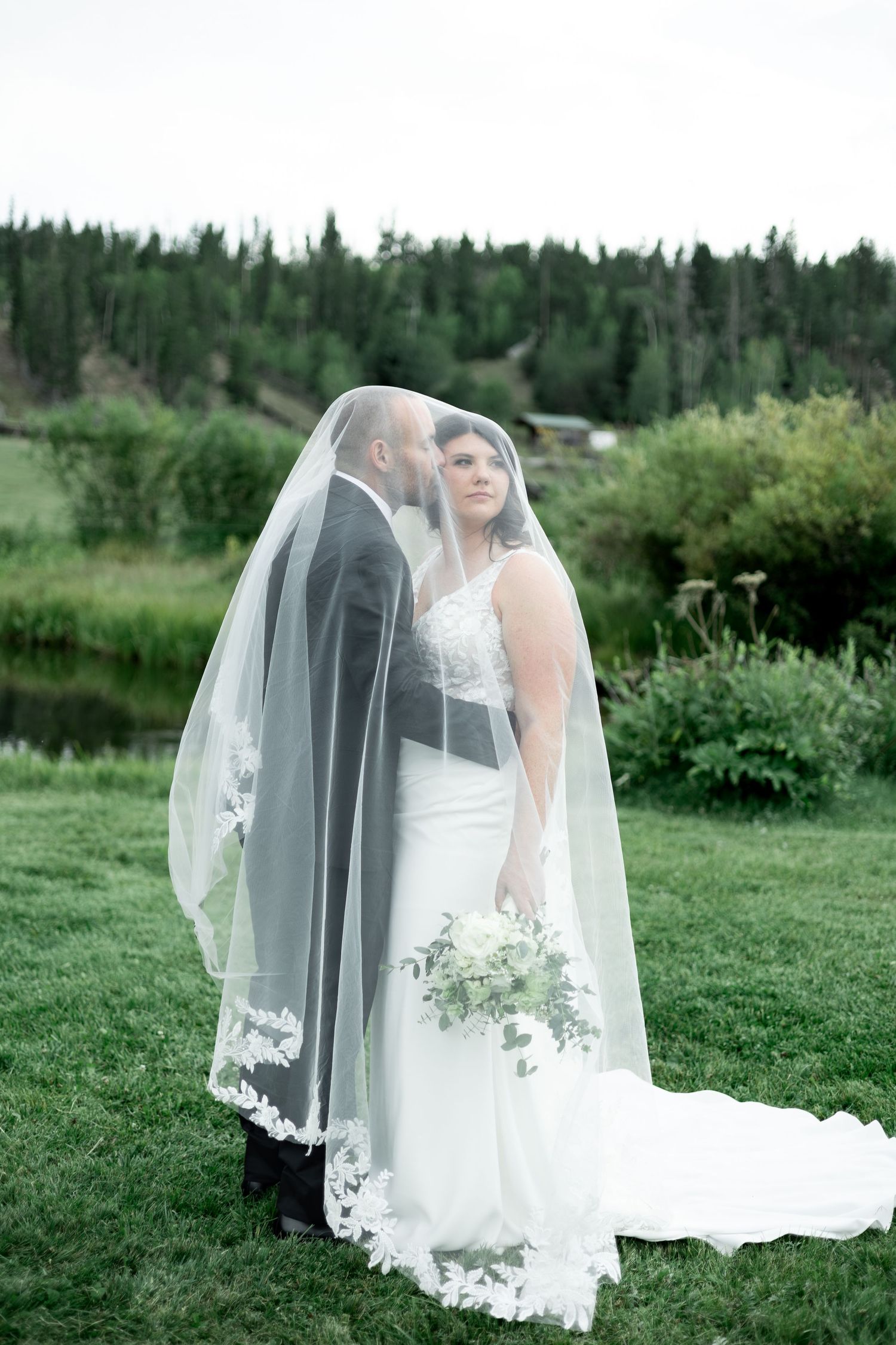 A romantic wedding couple poses together in a lush green outdoor setting with a flowing lace-trimmed veil.