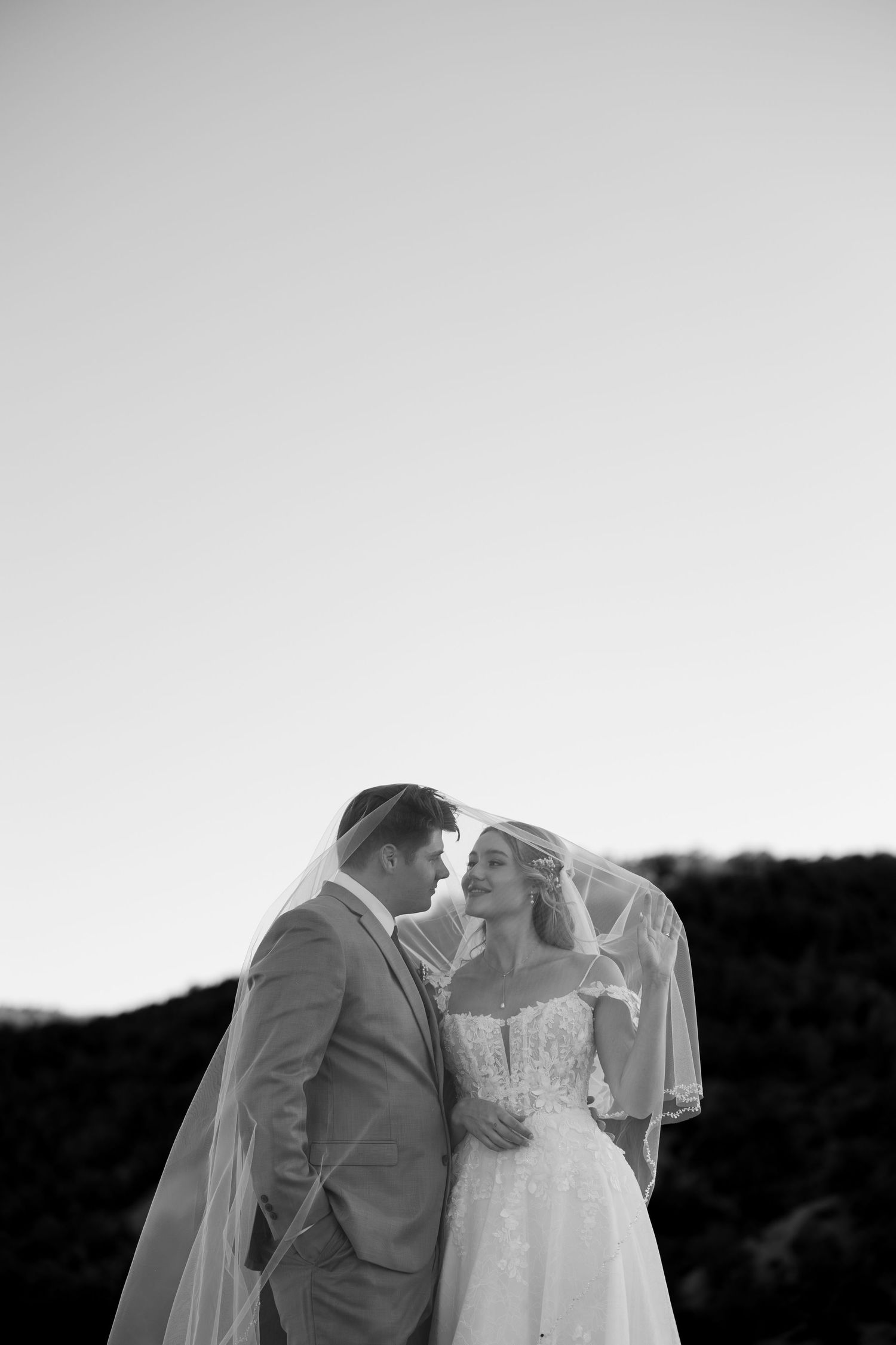 Black and white photo of bride and groom sharing romantic moment under wedding veil outdoors.