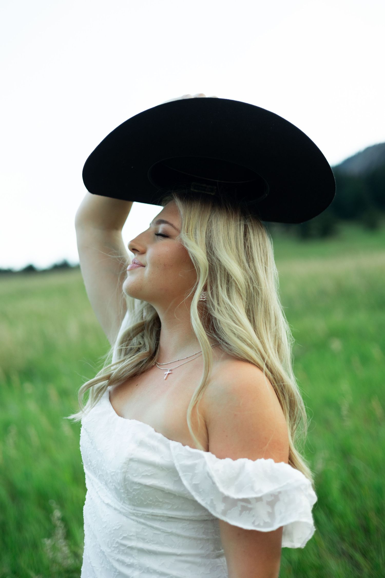 Someone wearing a black cowboy hat and white off-shoulder dress poses in a green field.