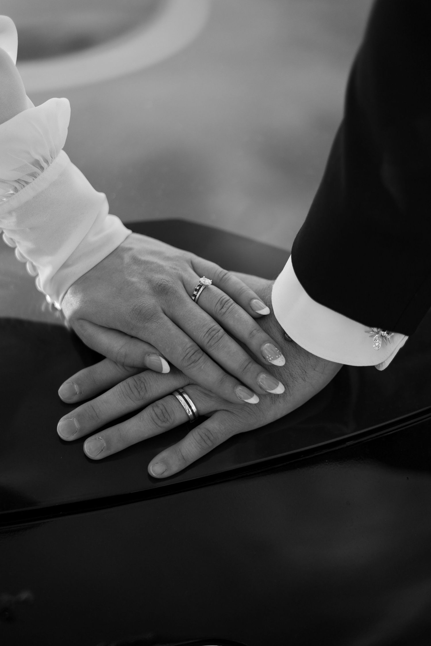 Black and white close-up photo of hands with wedding rings stacked together, showing white sleeve and formal attire.