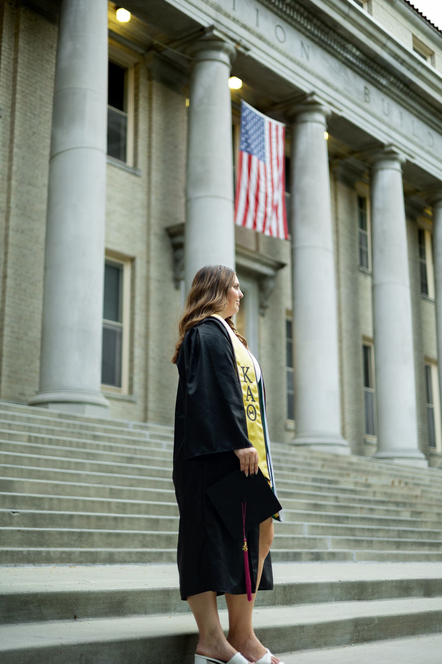 Graduate stands in black cap and gown in front of historic government building with American flag and columns.