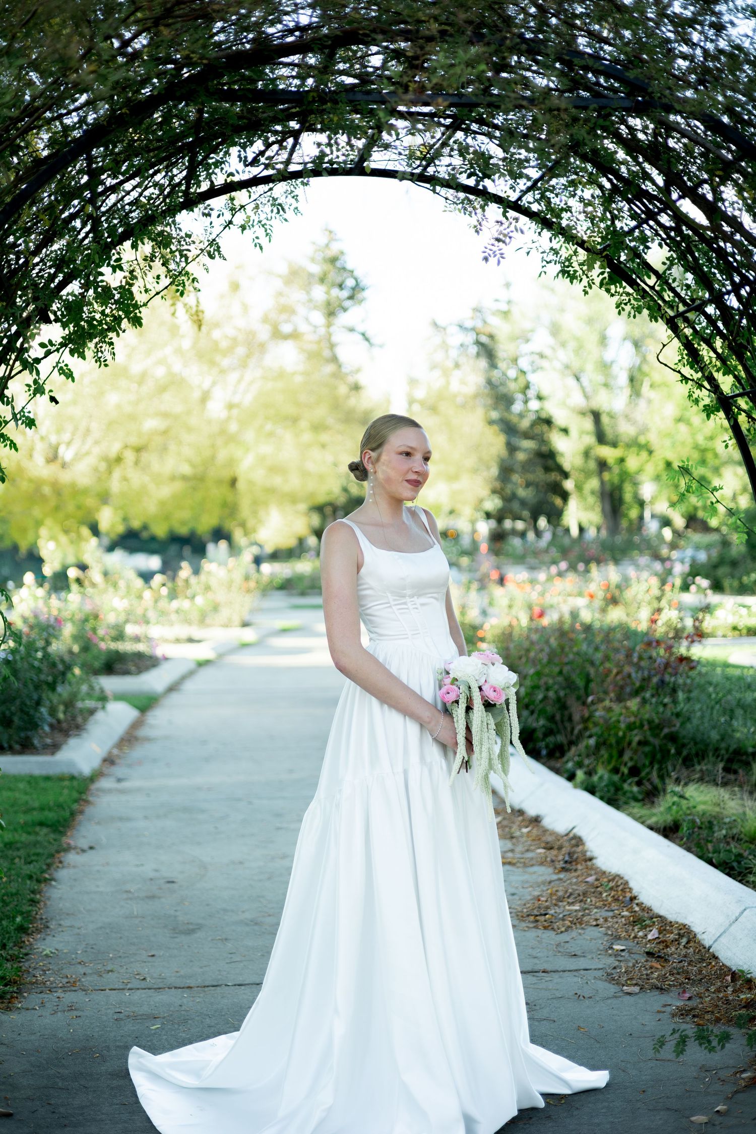 A figure in a flowing white wedding dress walks along a tree-lined path in a garden setting.