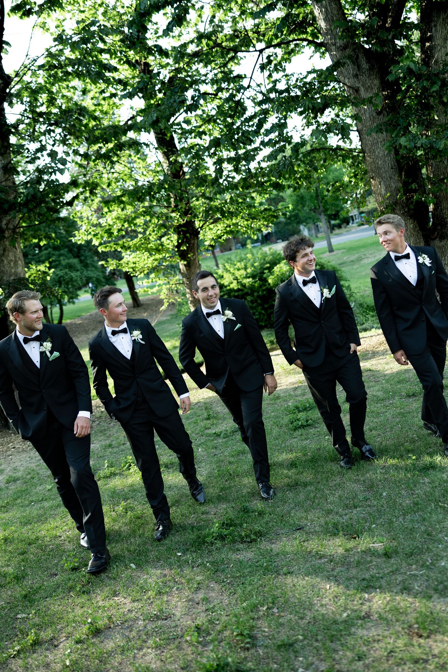 Five groomsmen in black tuxedos with bow ties walk together on green grass surrounded by lush trees.