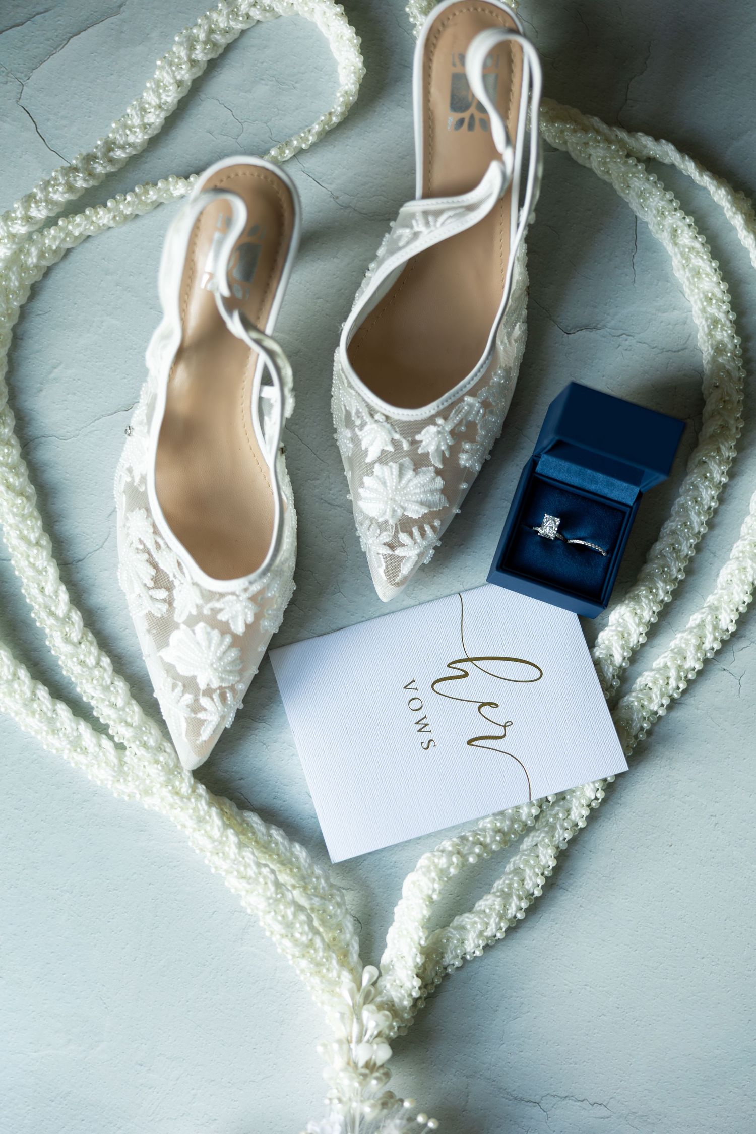Delicate white lace bridal shoes arranged with wedding stationery and a blue ring box.