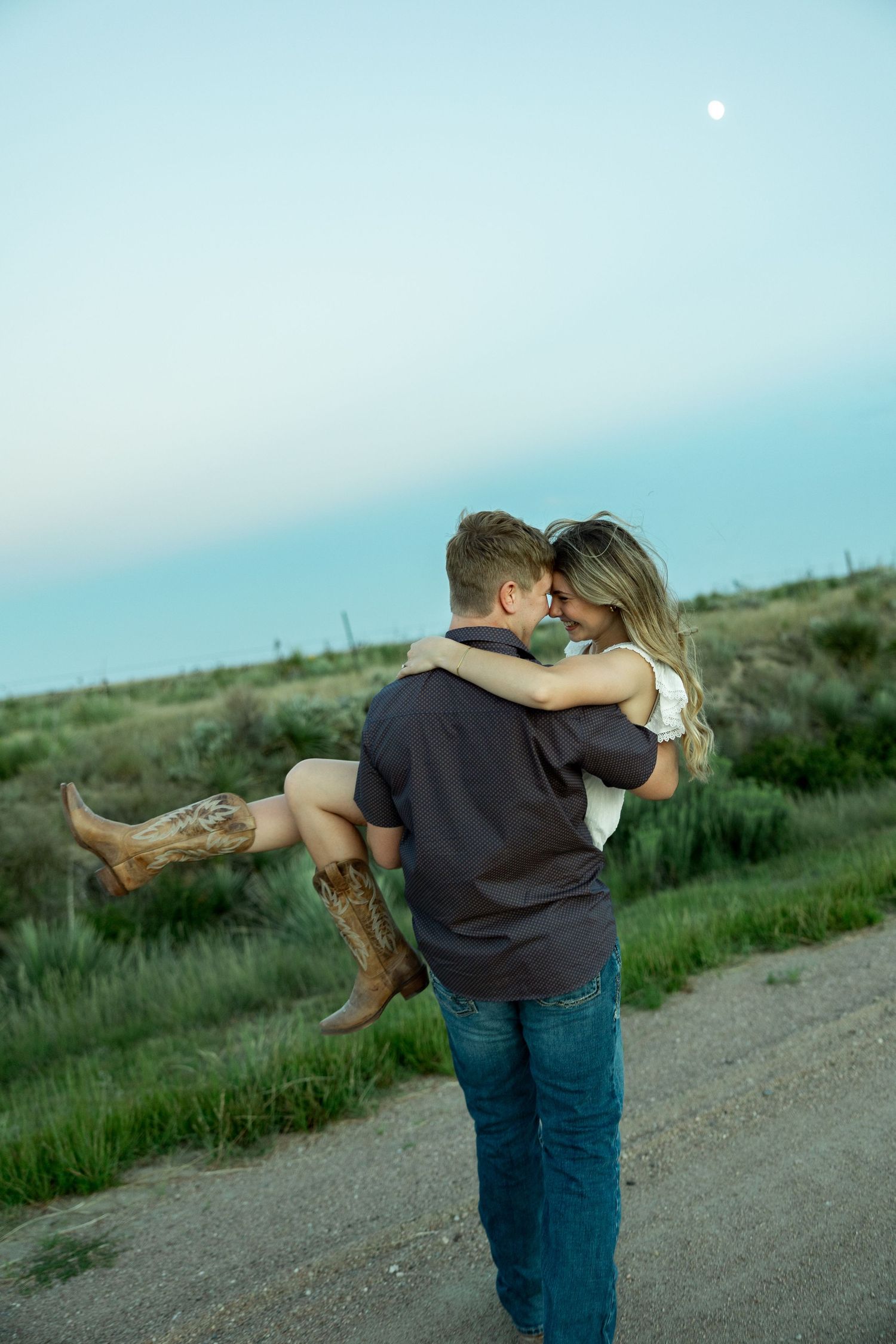 Man carrying woman in cowboy boots on a rural road at dusk with moon visible in the sky.