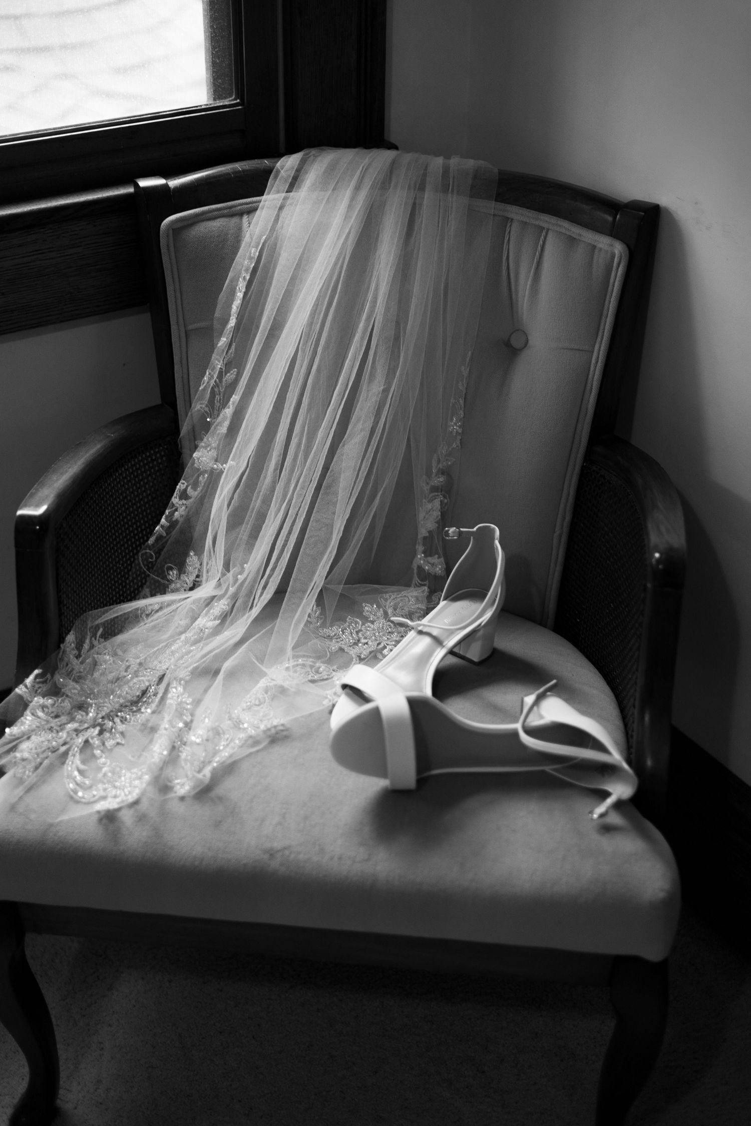 Black and white photo of bridal veil and white heels on vintage chair near window.