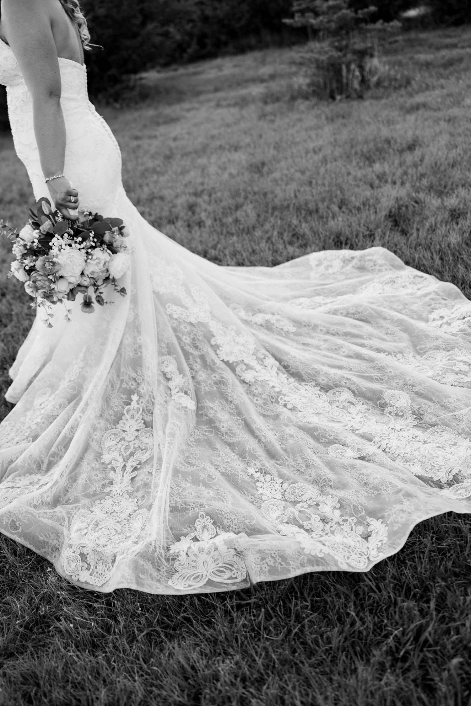 Black and white photo of a bride in a lace mermaid wedding gown with a long train, holding a floral bouquet.