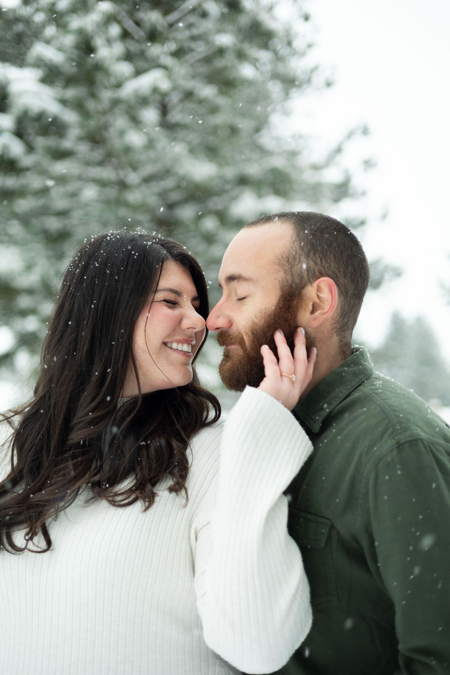 A candid winter moment captured between a couple in white and green against snowy evergreens.