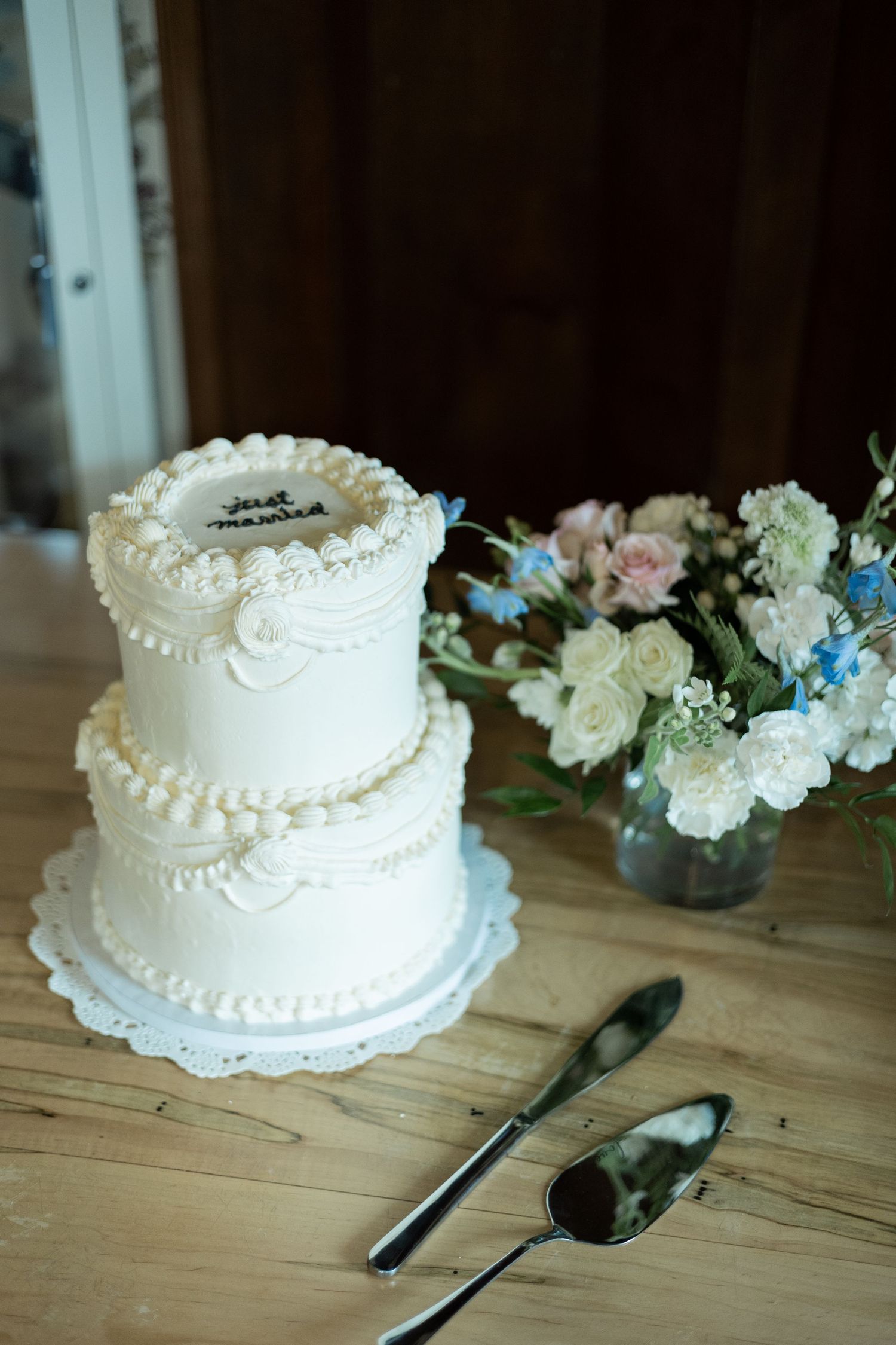 A white two-tiered wedding cake with pearl decorations sits on a wooden table next to floral arrangements.