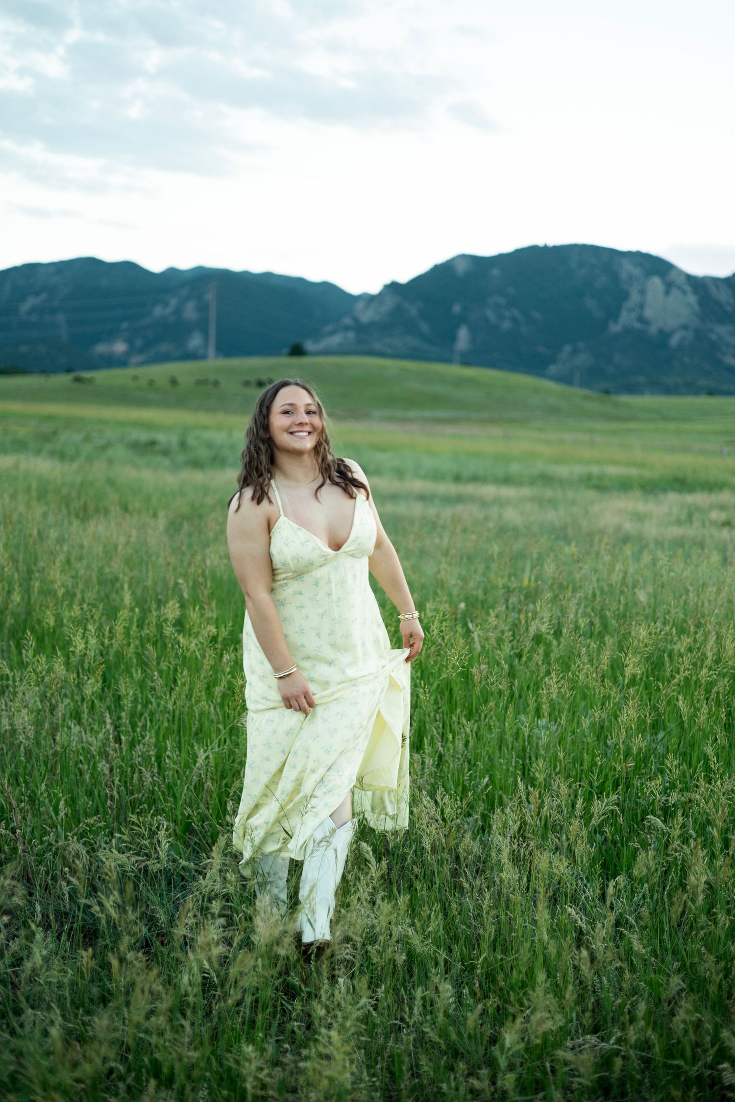 A person in a flowing cream dress walks through a green meadow with mountains in the background.