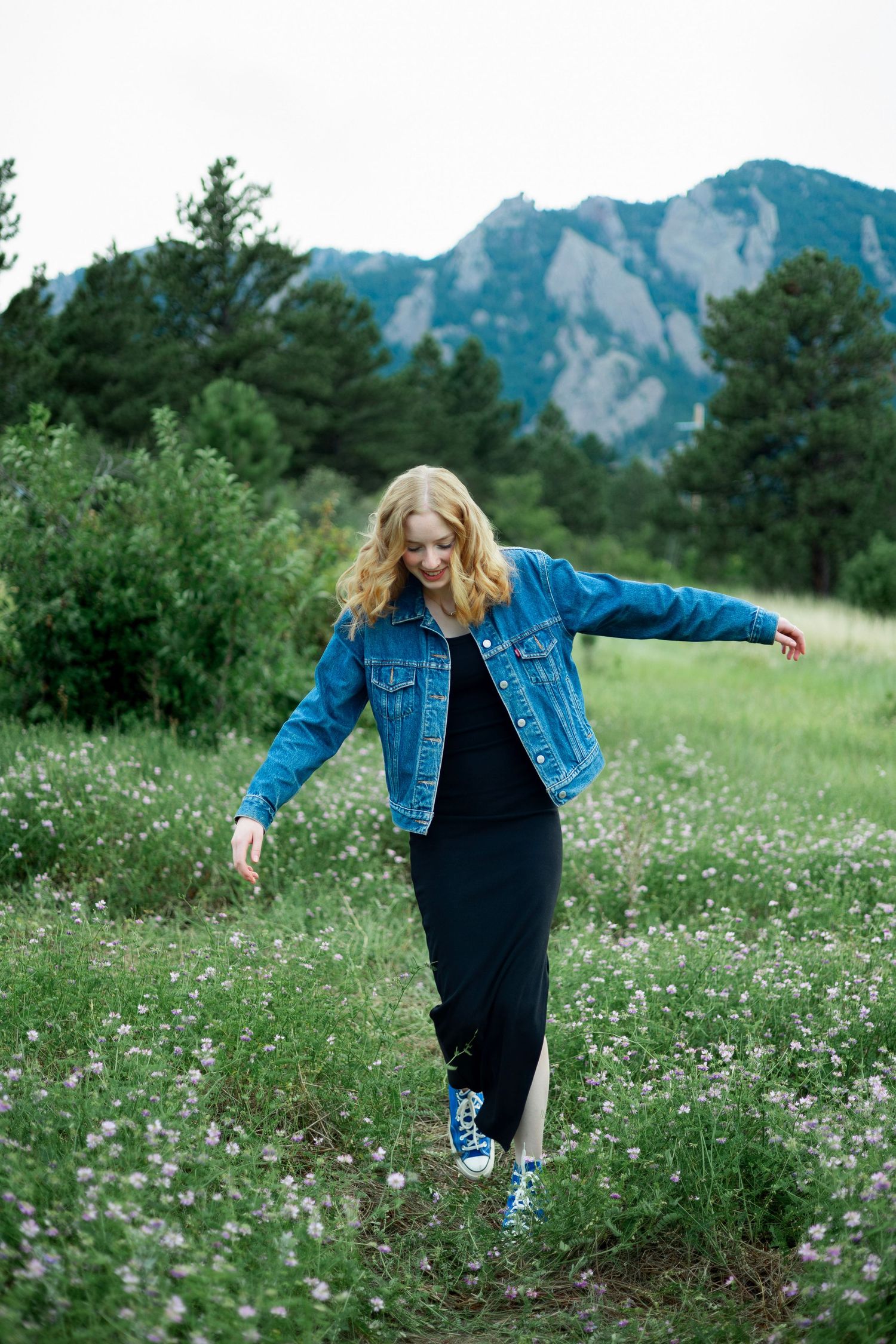 Woman in denim jacket and black dress walking through wildflower meadow with Rocky Mountains in background.