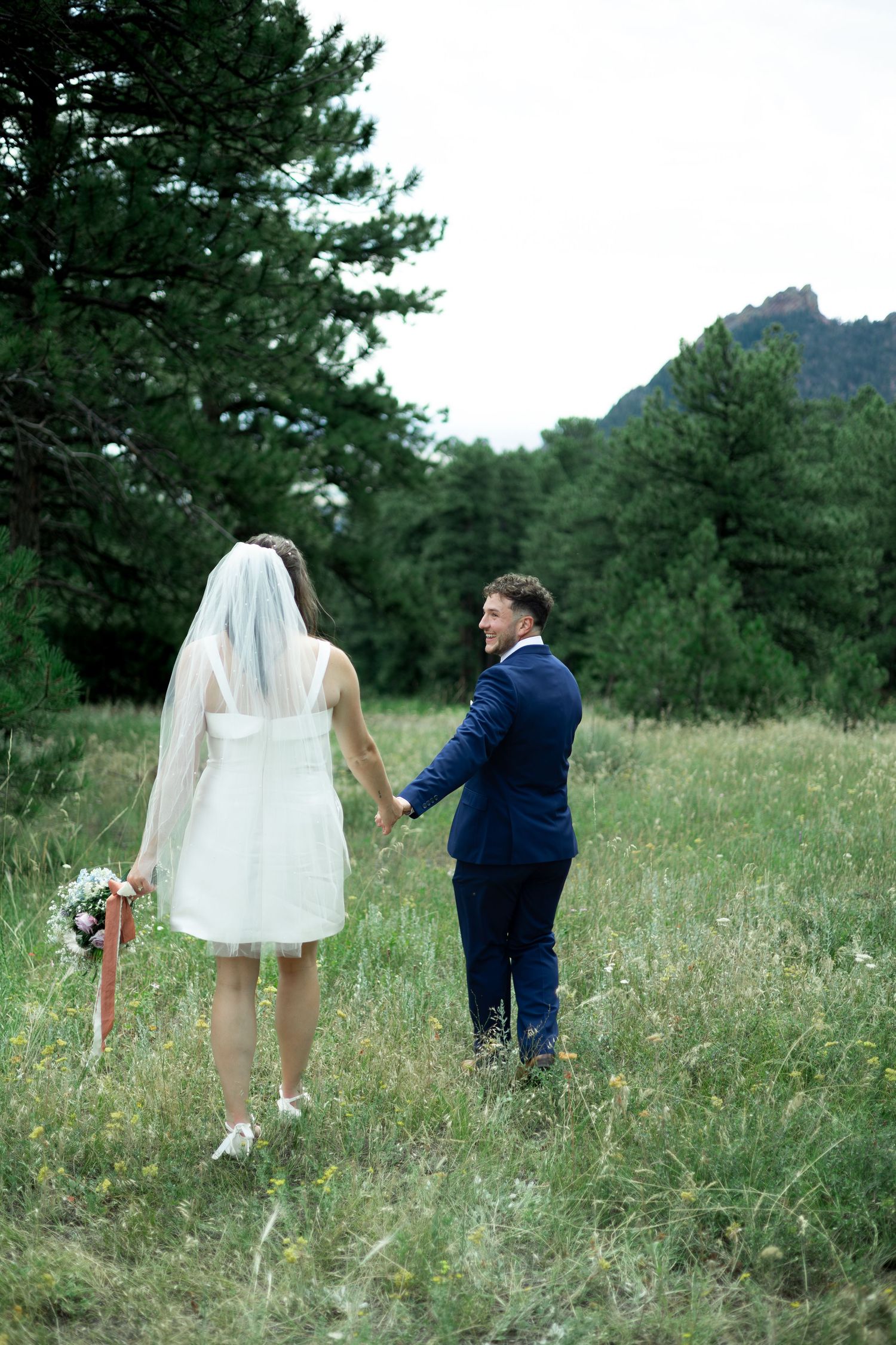Bride in short white dress and veil holds hands with groom in navy suit walking through green meadow.