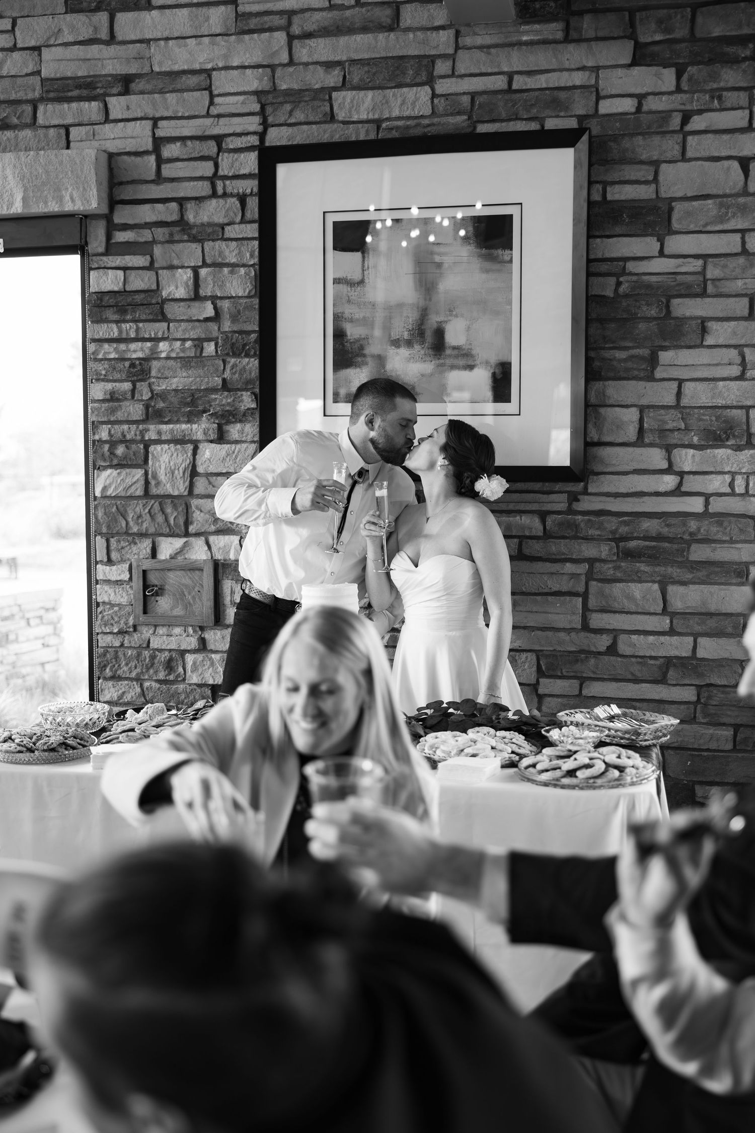 Bride and groom share a kiss at wedding reception near food table with guests raising glasses in black and white photo.