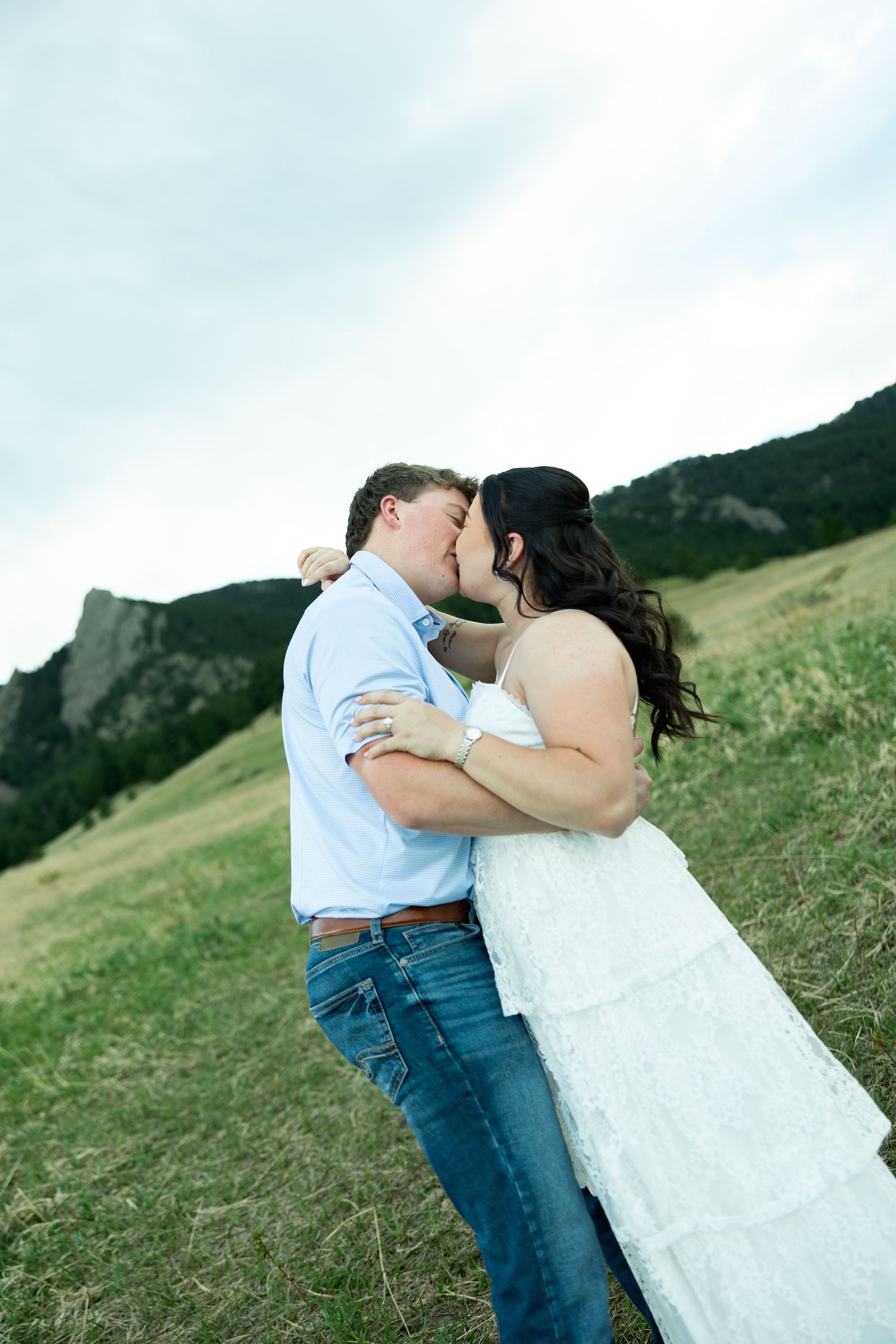 Couple sharing a romantic kiss in a green meadow with rocky mountain backdrop, woman in white dress and man in blue shirt.