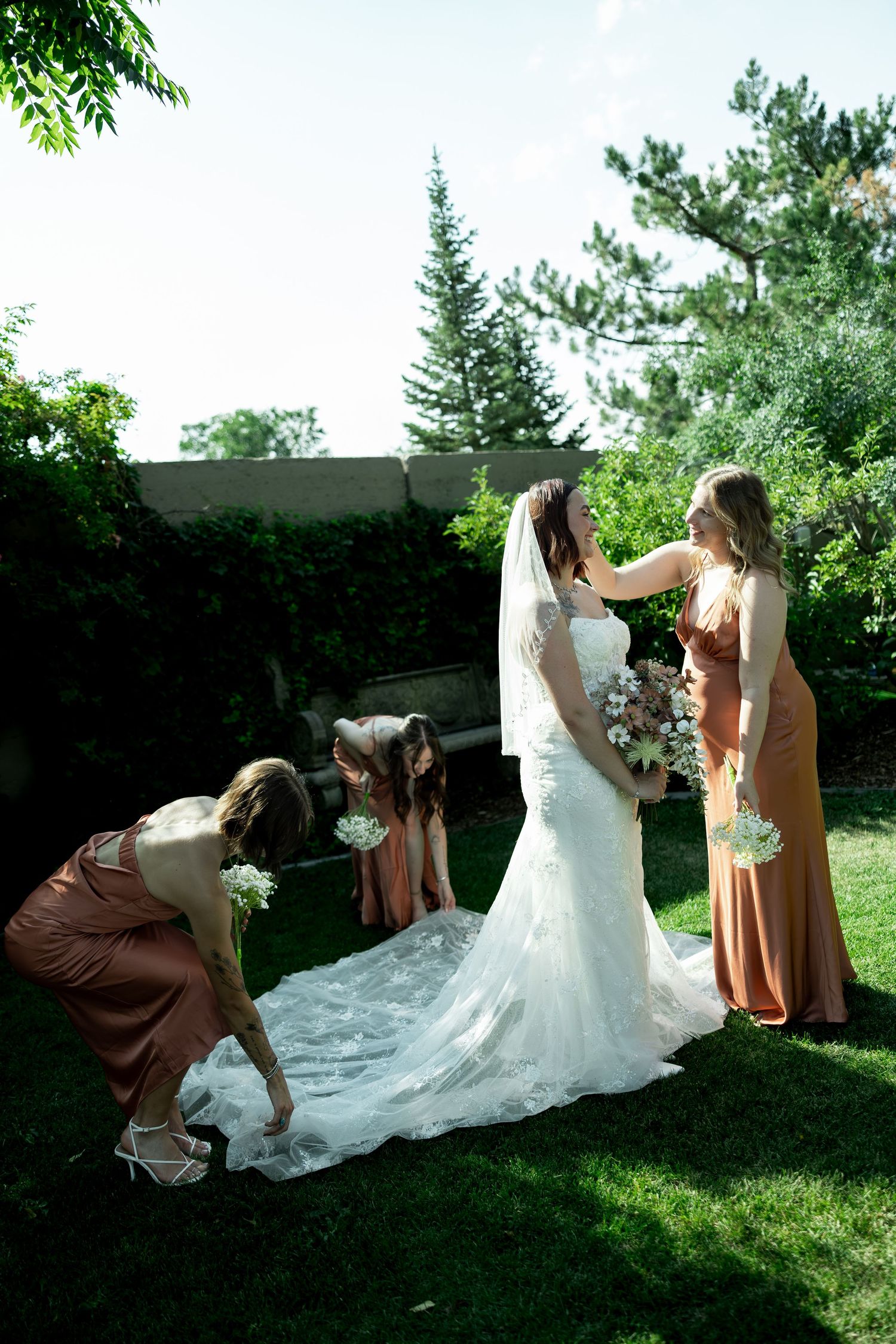 Bridesmaids in rust dresses help bride arrange her lace wedding gown train outdoors in a lush green garden.
