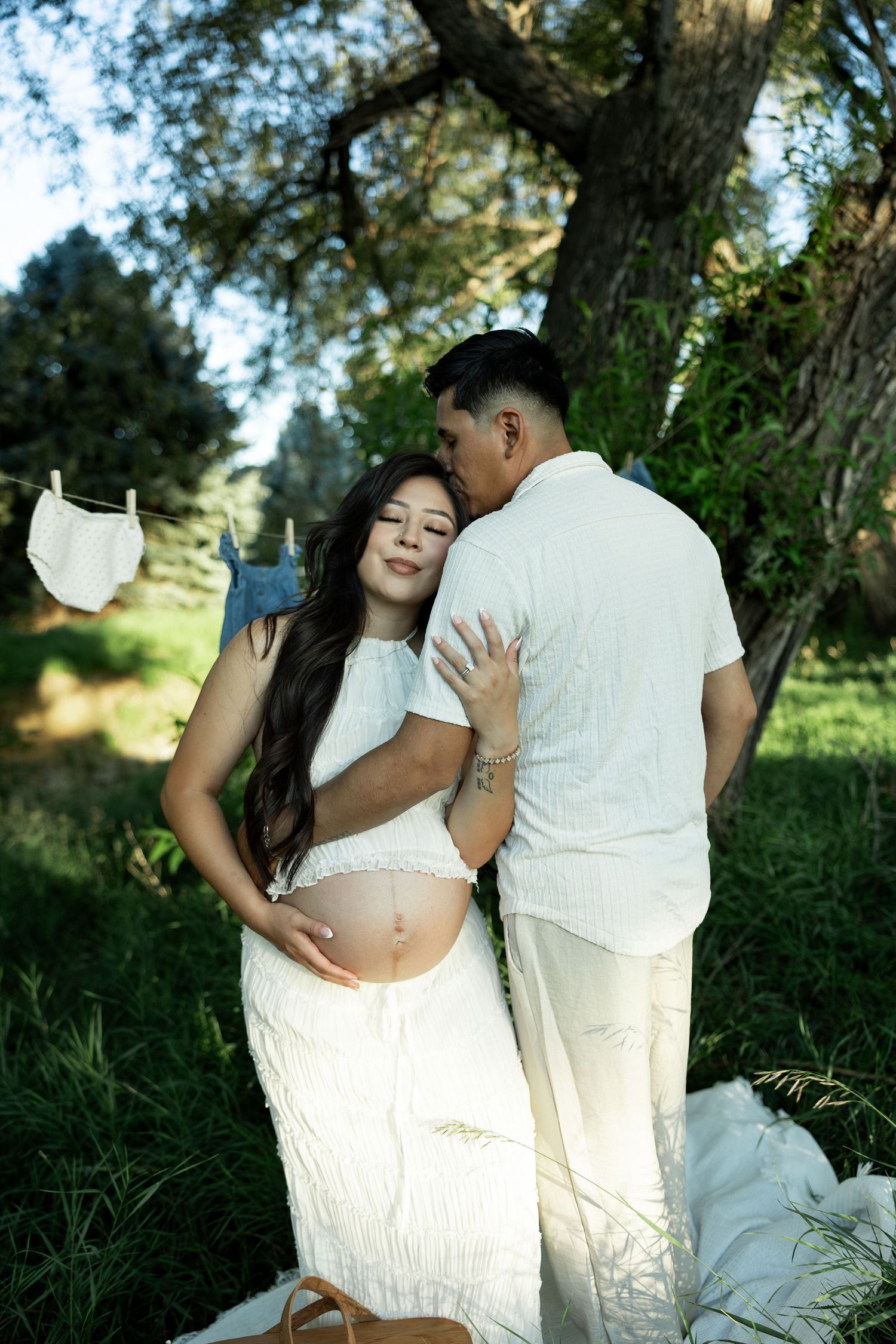 Pregnant couple in white outfits embracing outdoors under trees with baby clothes hanging on a line.