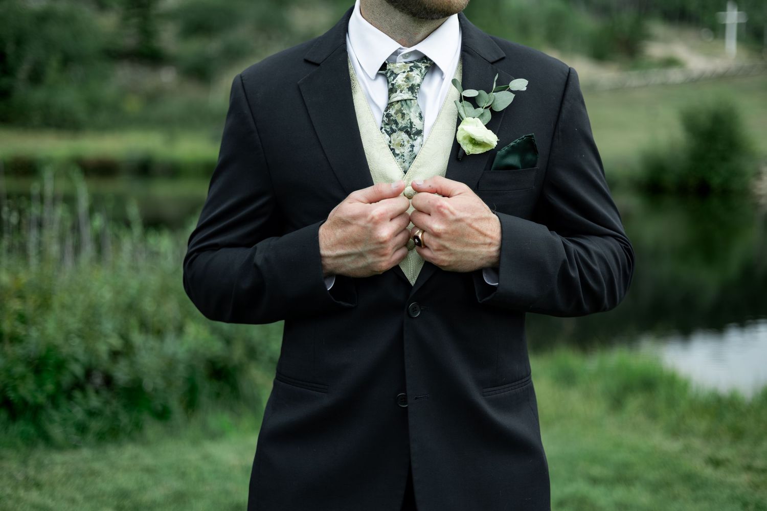 A groom in a black tuxedo adjusts his white boutonniere while standing outdoors in a garden setting.