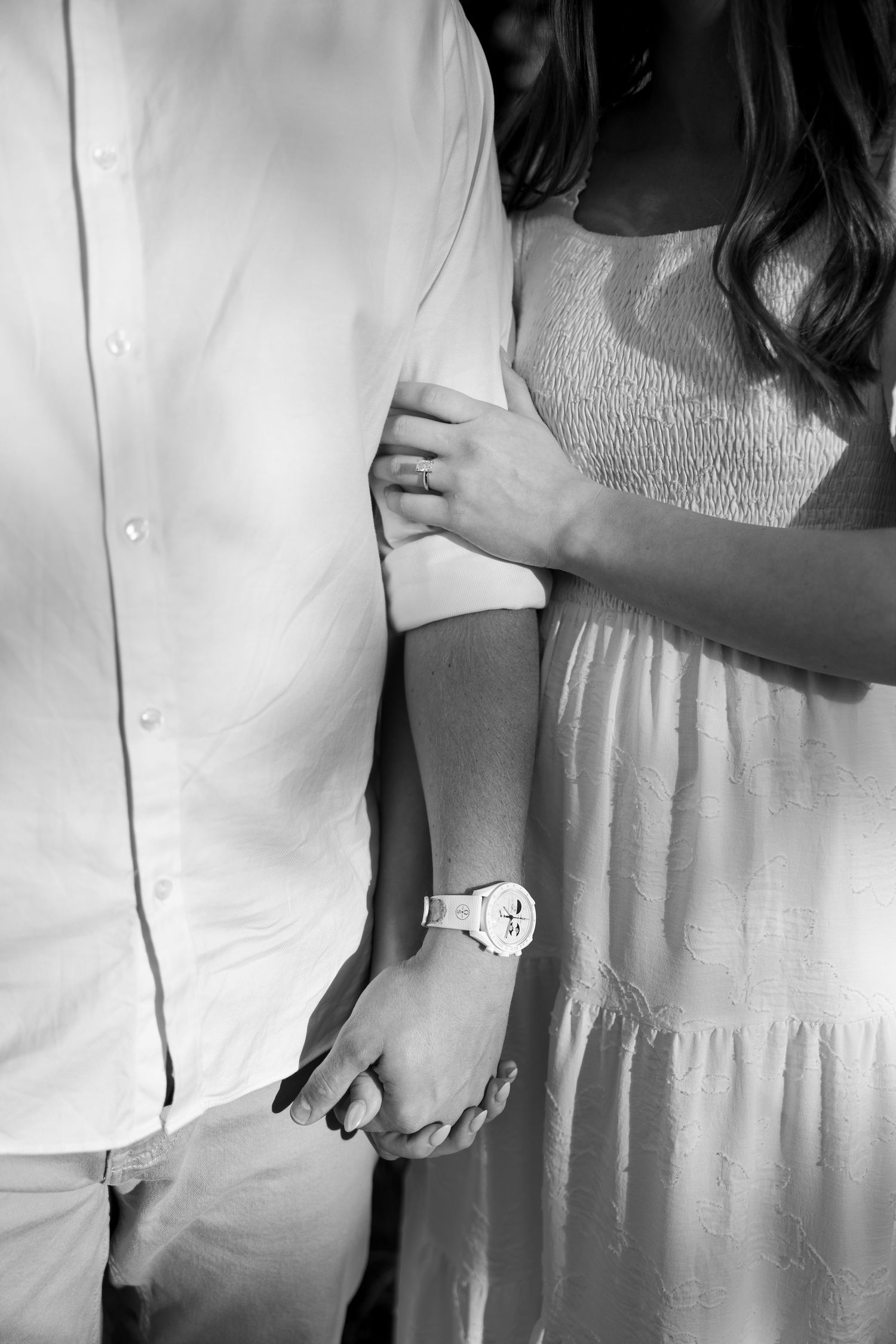 Black and white photo of a couple holding hands, woman in white dress with watch, man in white shirt.