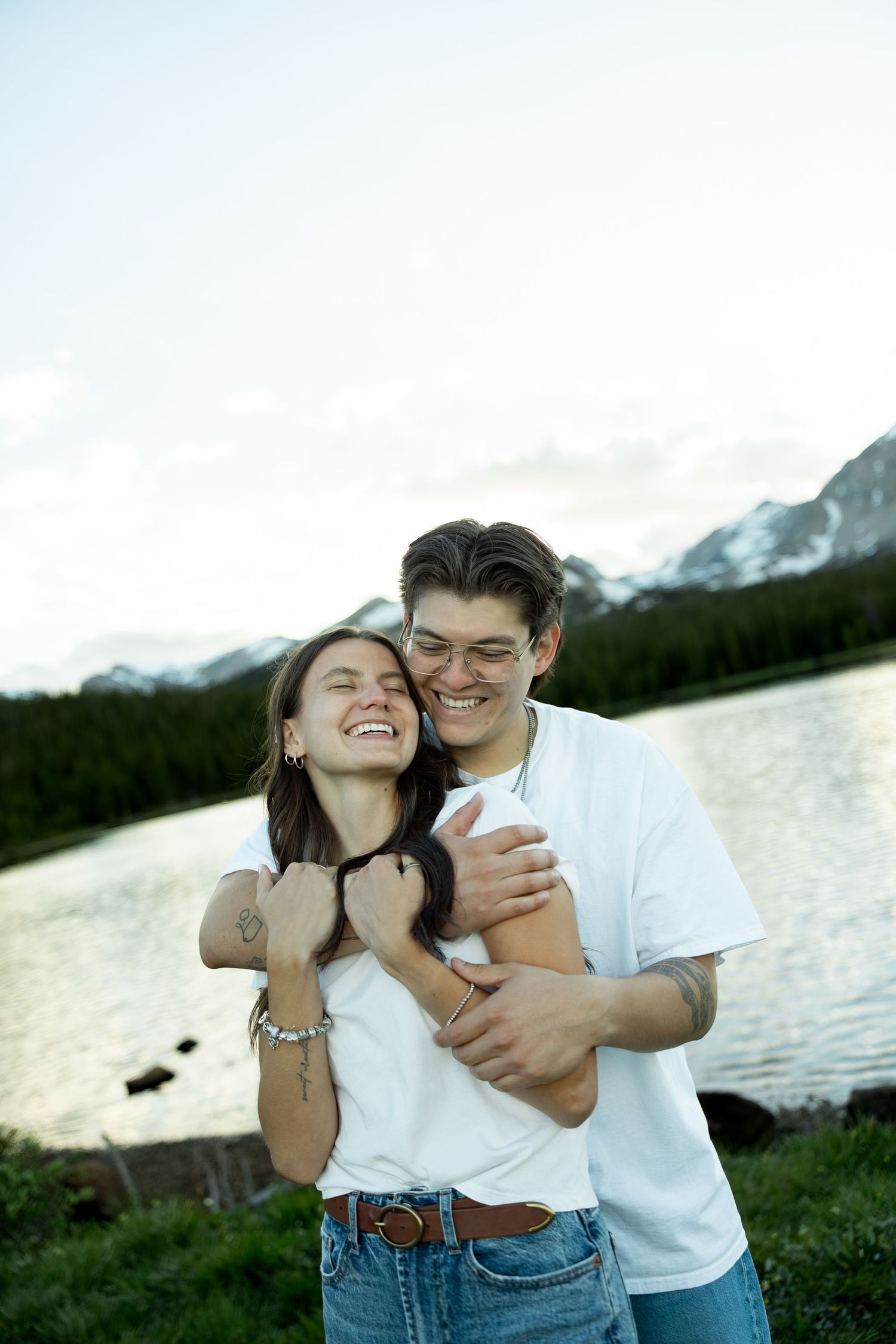 Happy couple embracing and laughing by a mountain lake with snow-capped peaks and forest in the background.
