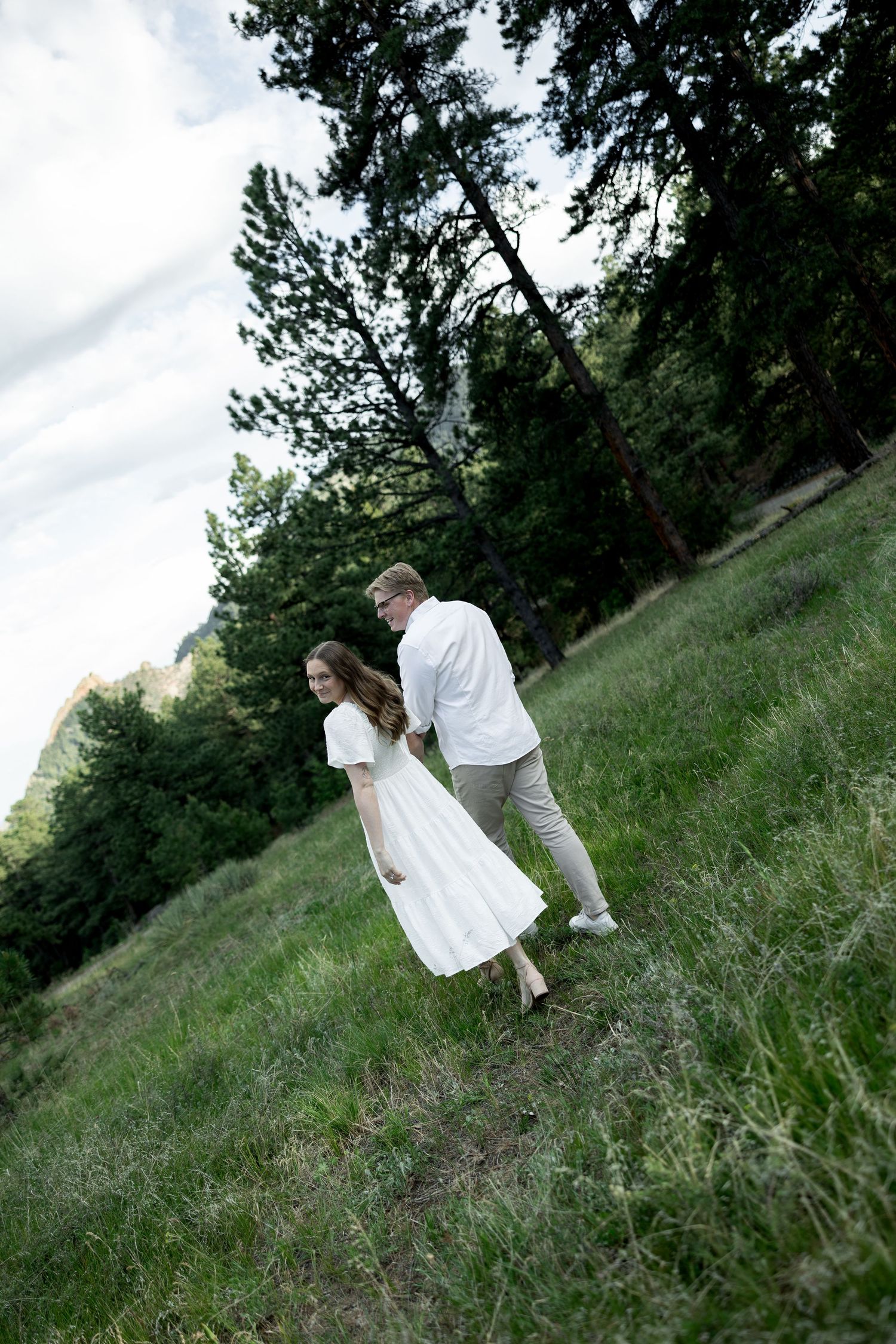 Couple in white outfits walking through a lush green meadow surrounded by tall trees in a scenic outdoor setting.