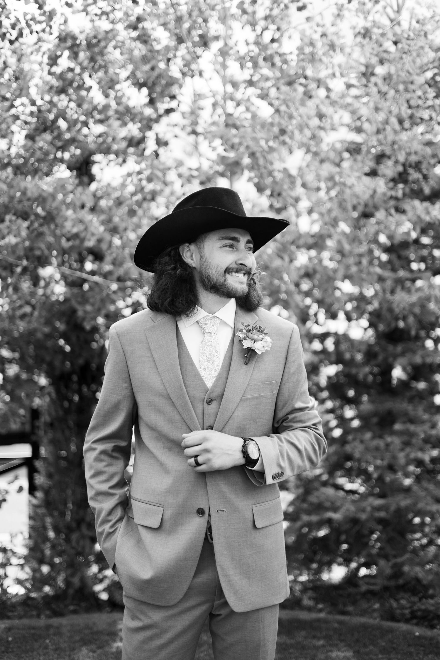 Portrait of a groom in a grey suit and black cowboy hat smiling outdoors.