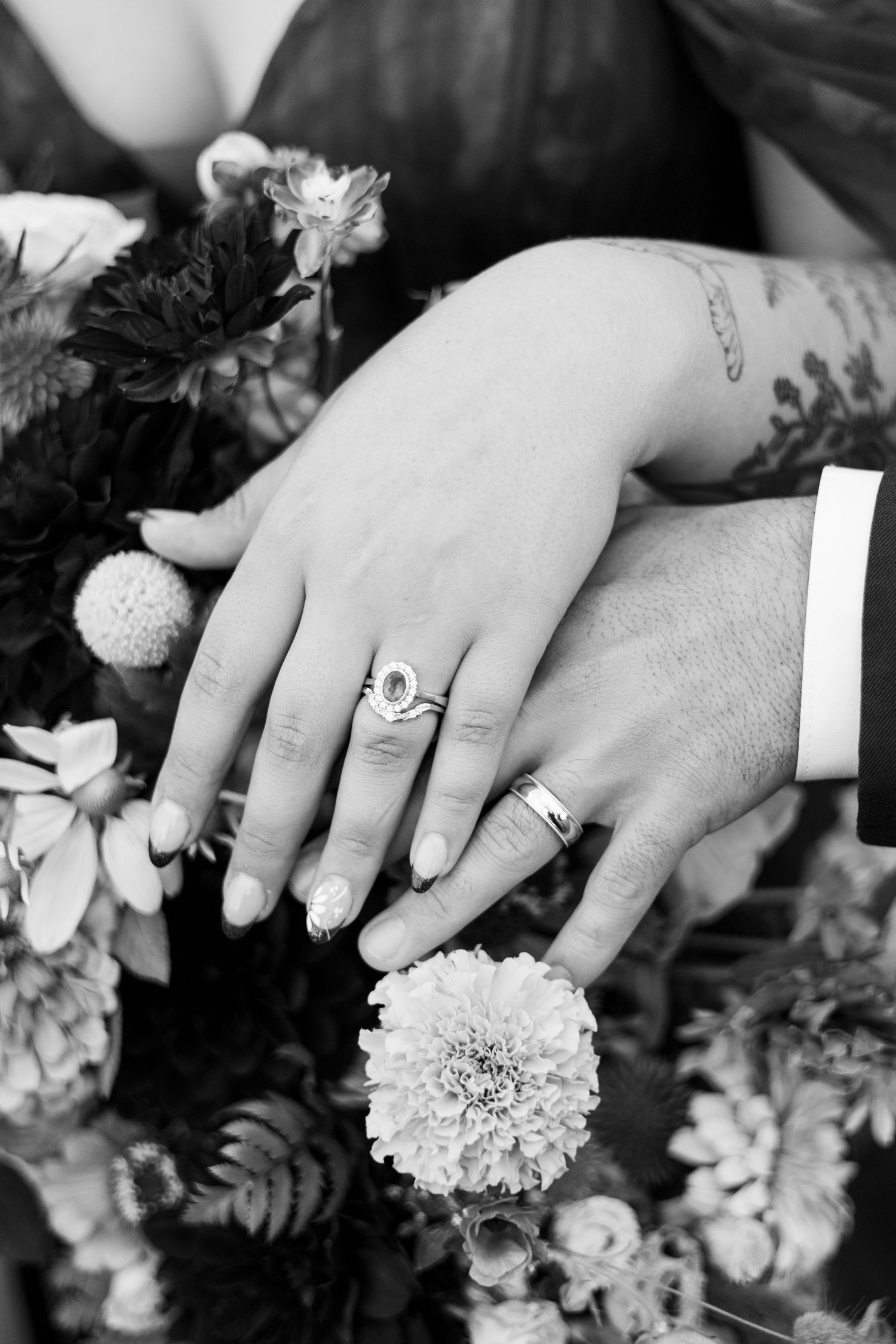 Close-up of wedding rings and hands resting on a floral bouquet in black and white.