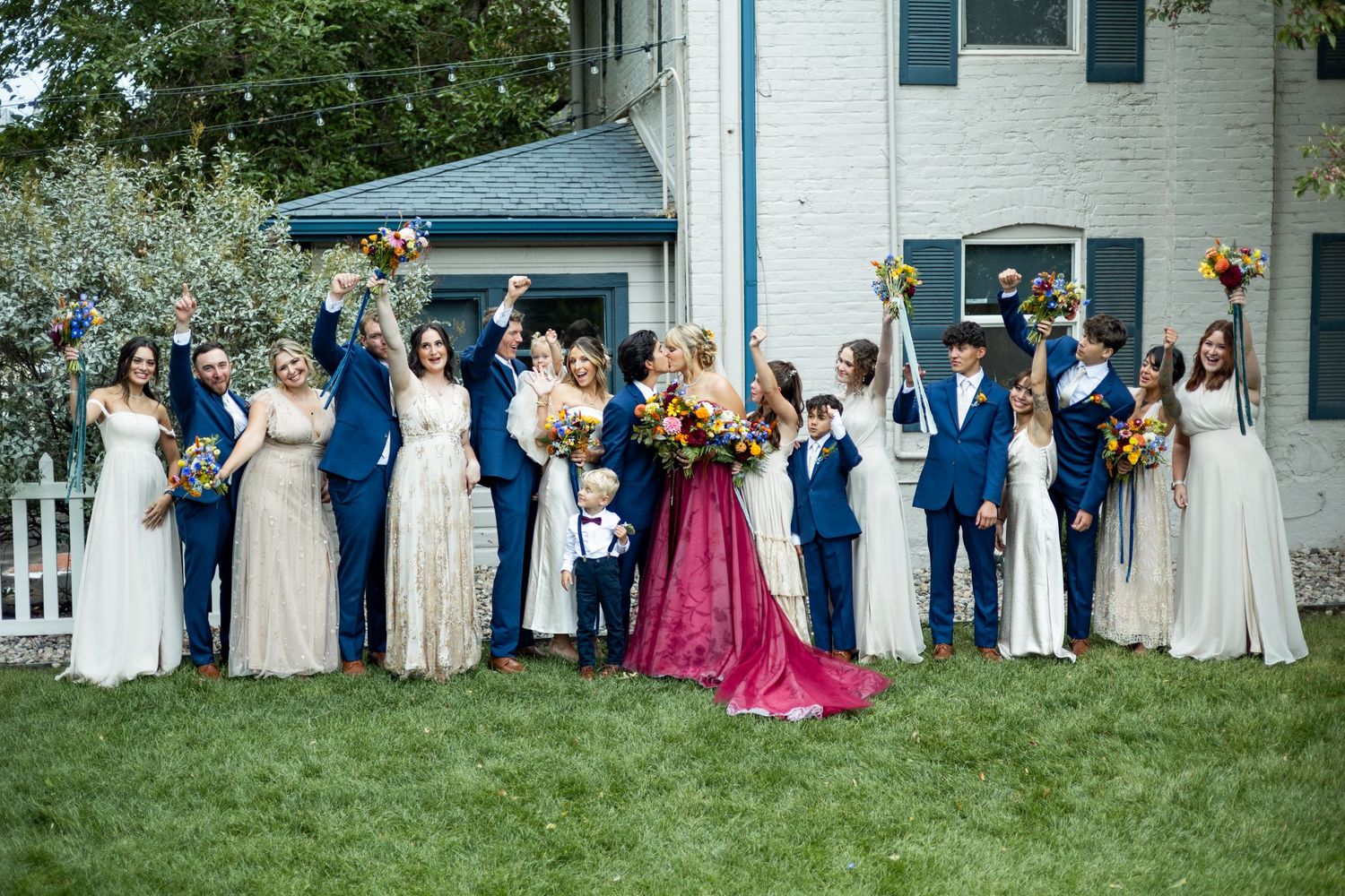 Wedding party in blue and white dresses poses on lawn outside white building with burgundy accent dress.