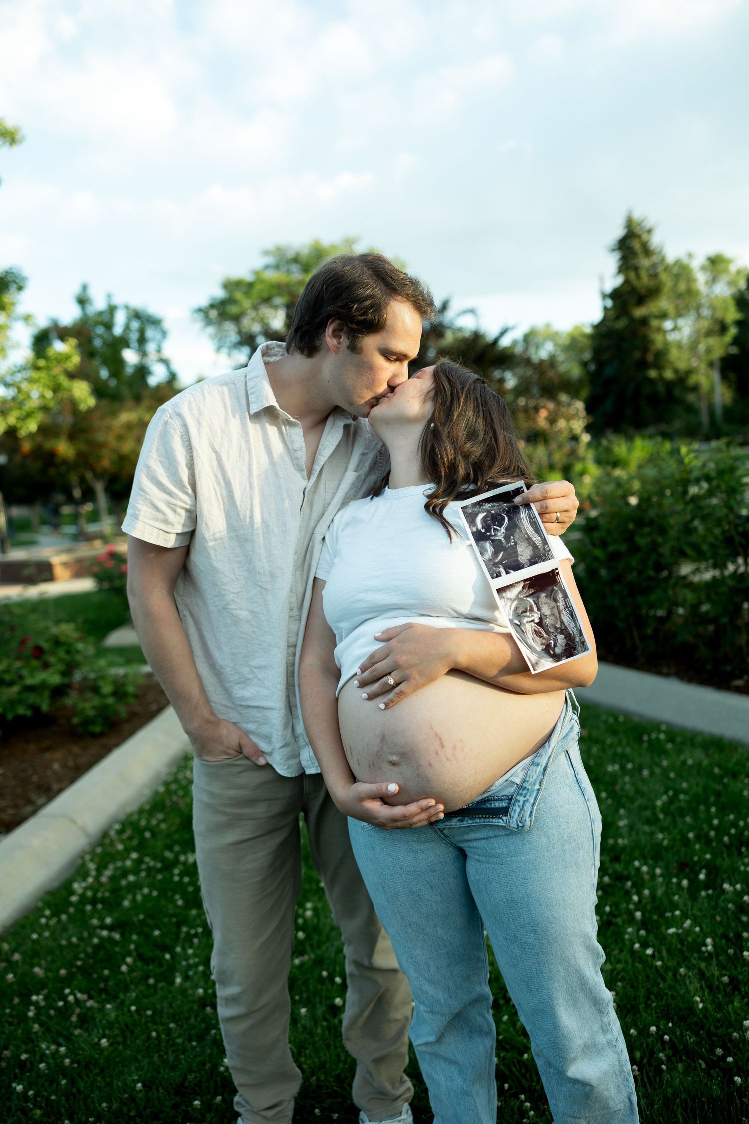 Couple kissing outdoors while pregnant woman holds ultrasound photos and cradles baby bump in lush green park.