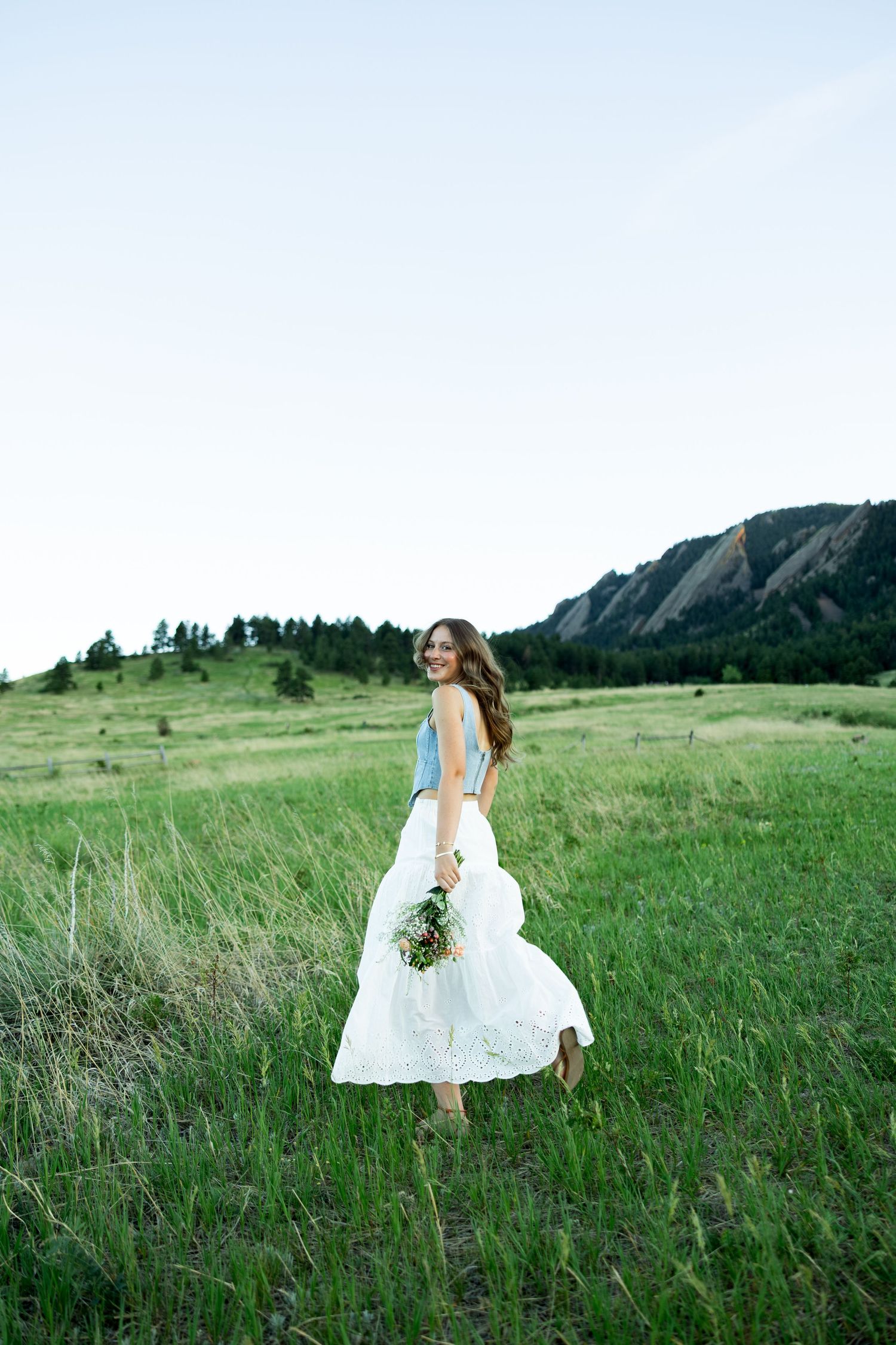 Bride twirling in white skirt and blue top holding bouquet in green field with mountain backdrop.