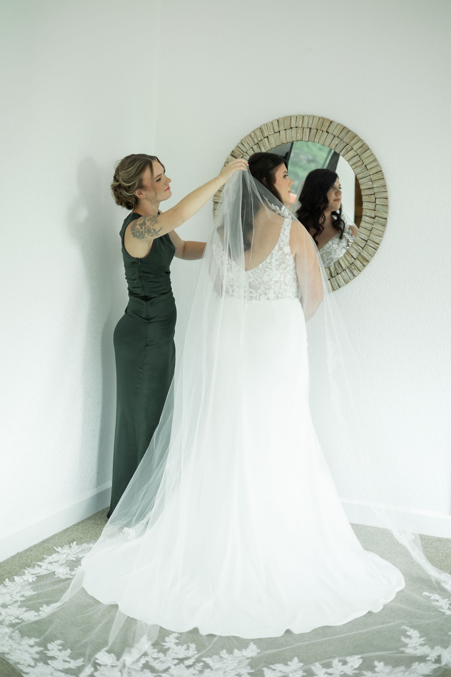 Two figures in contrasting green and white dresses pose dramatically against a light wall with a circular mirror.