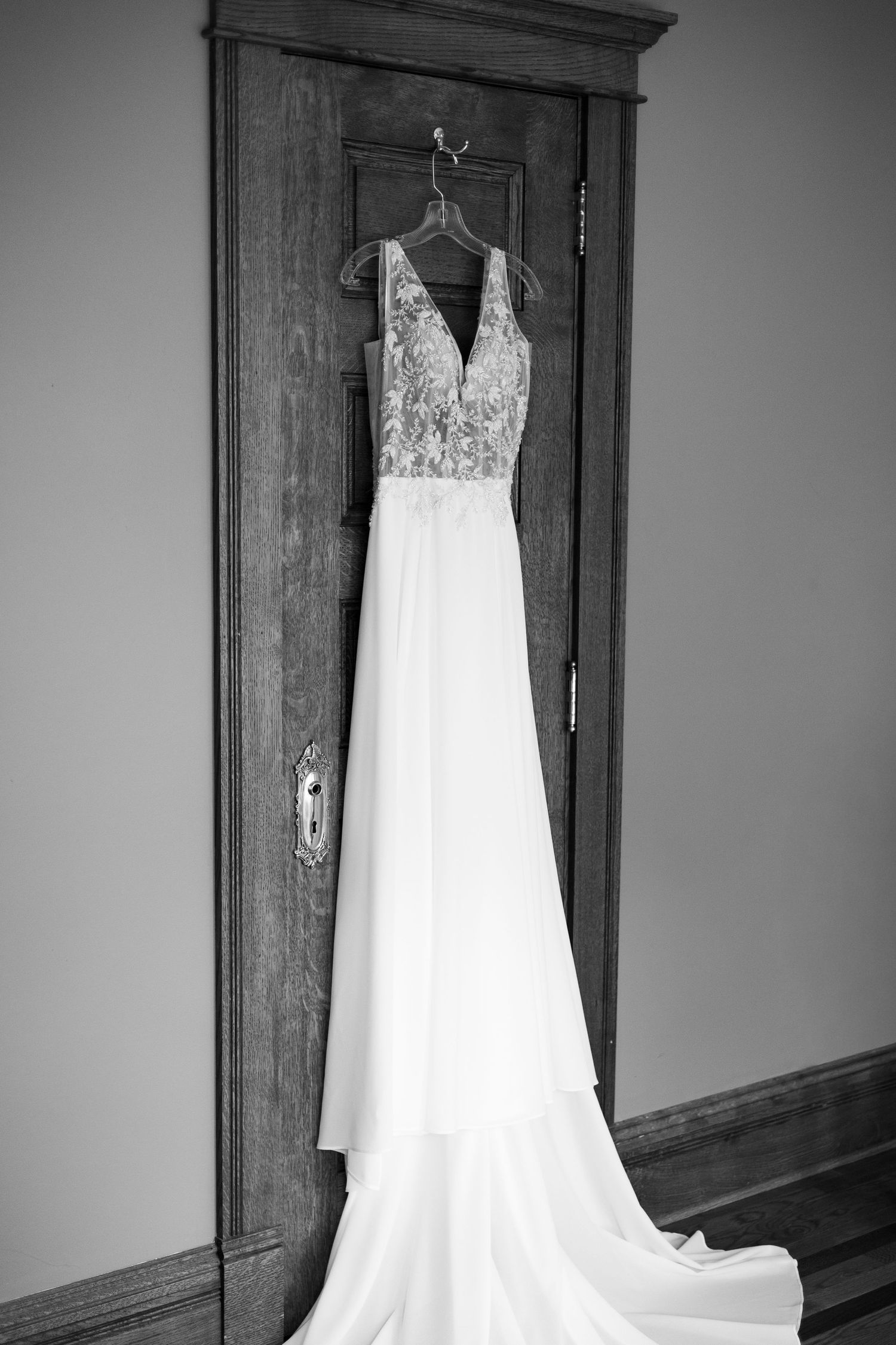 An elegant white lace wedding dress hangs on a dark wooden door frame against a minimalist grey wall background.