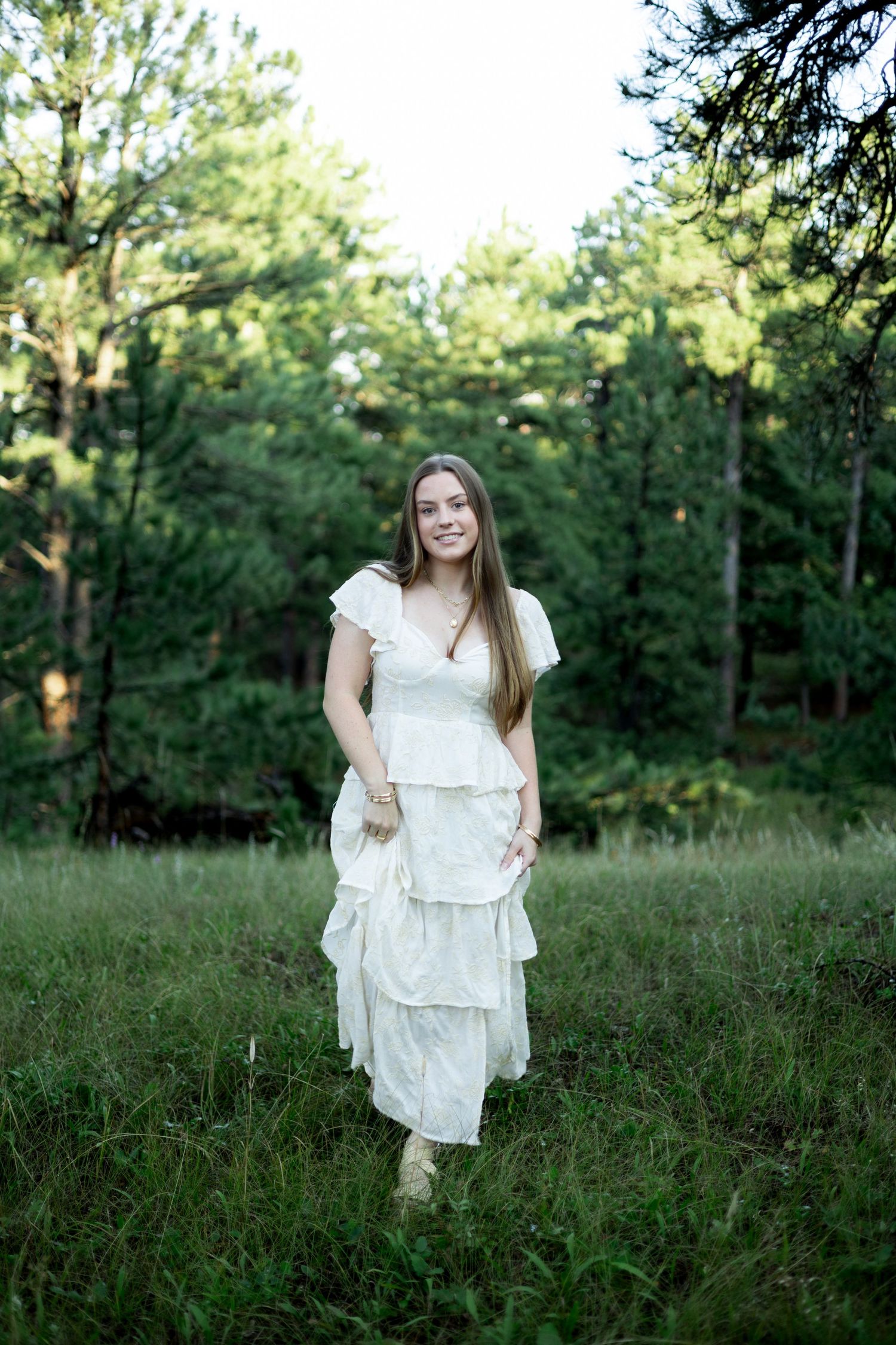 Young woman in white tiered ruffle dress walking through lush green forest meadow, smiling naturally.