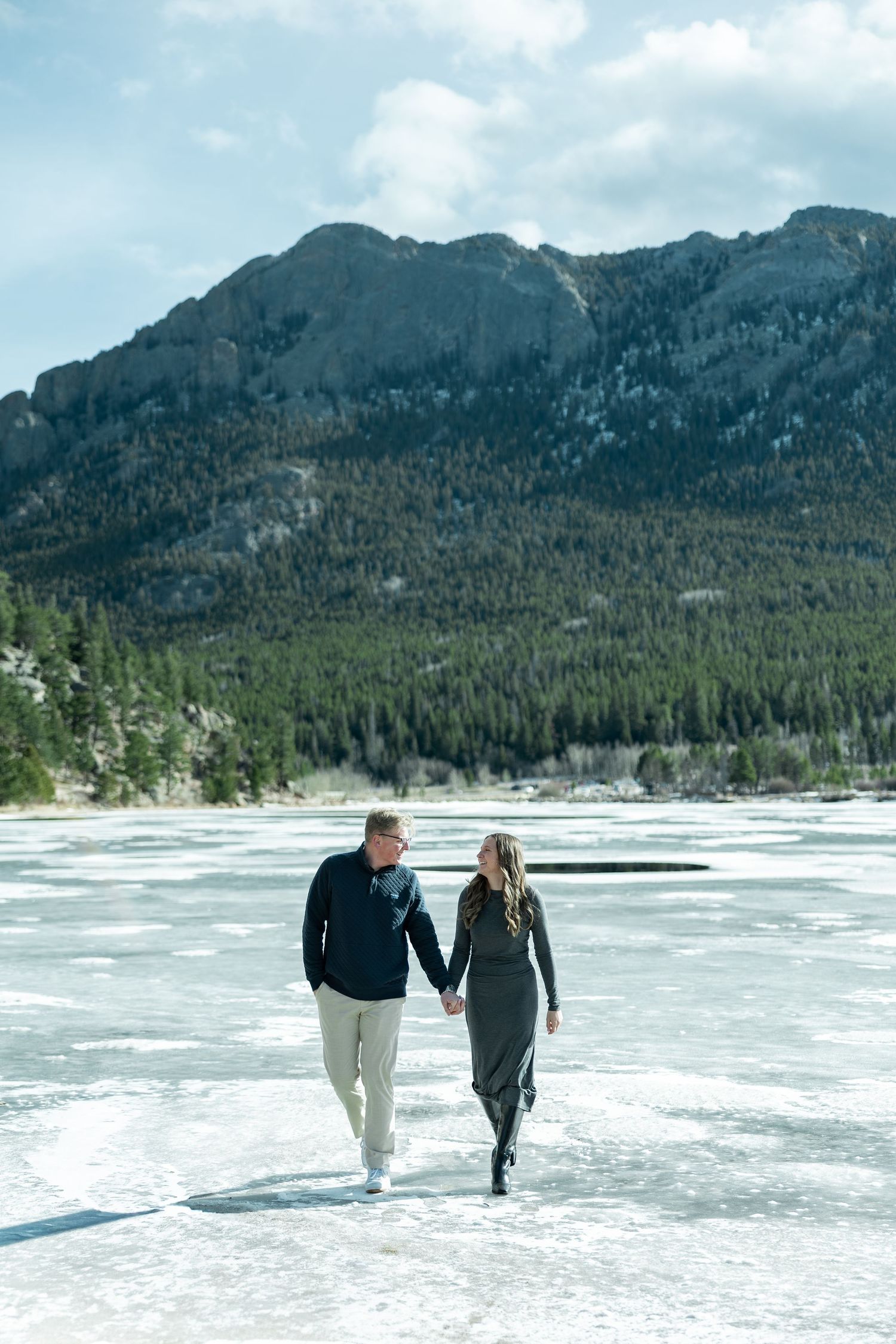 Couple holding hands walking on frozen lake with mountain and pine forest backdrop in winter.