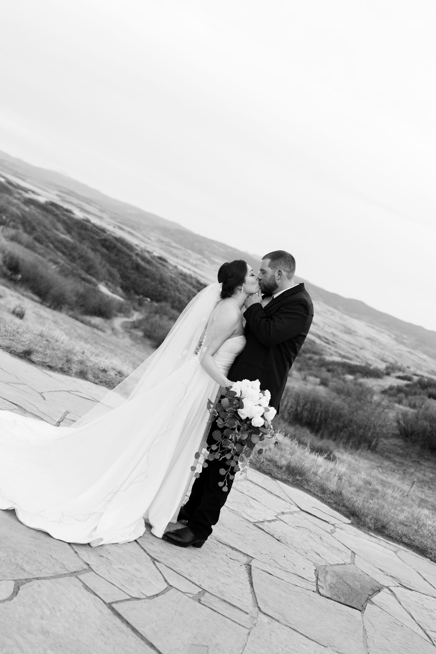 Black and white photo of bride and groom kissing on stone terrace with bouquet and flowing veil in wind.