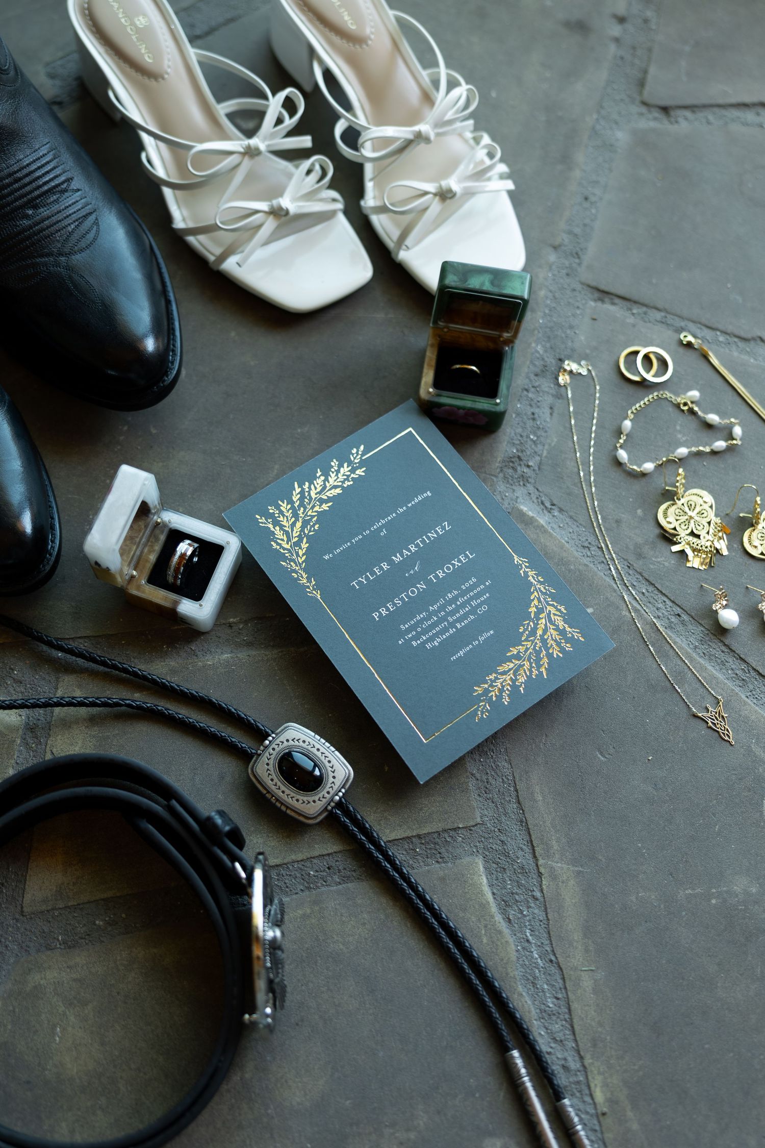 Elegant wedding flat lay featuring navy invitation, gold jewelry, white heels, black dress shoes, and a bolo tie on stone.
