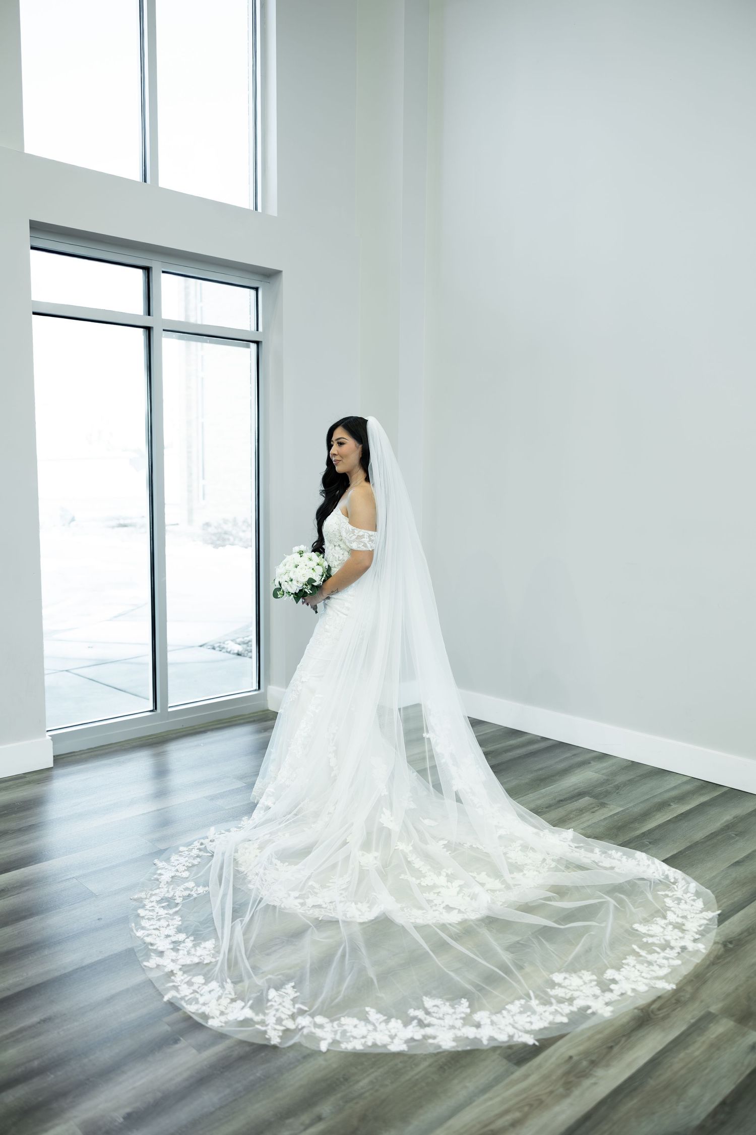 A bride in an elegant white gown with a dramatic cathedral-length lace veil stands by a window.
