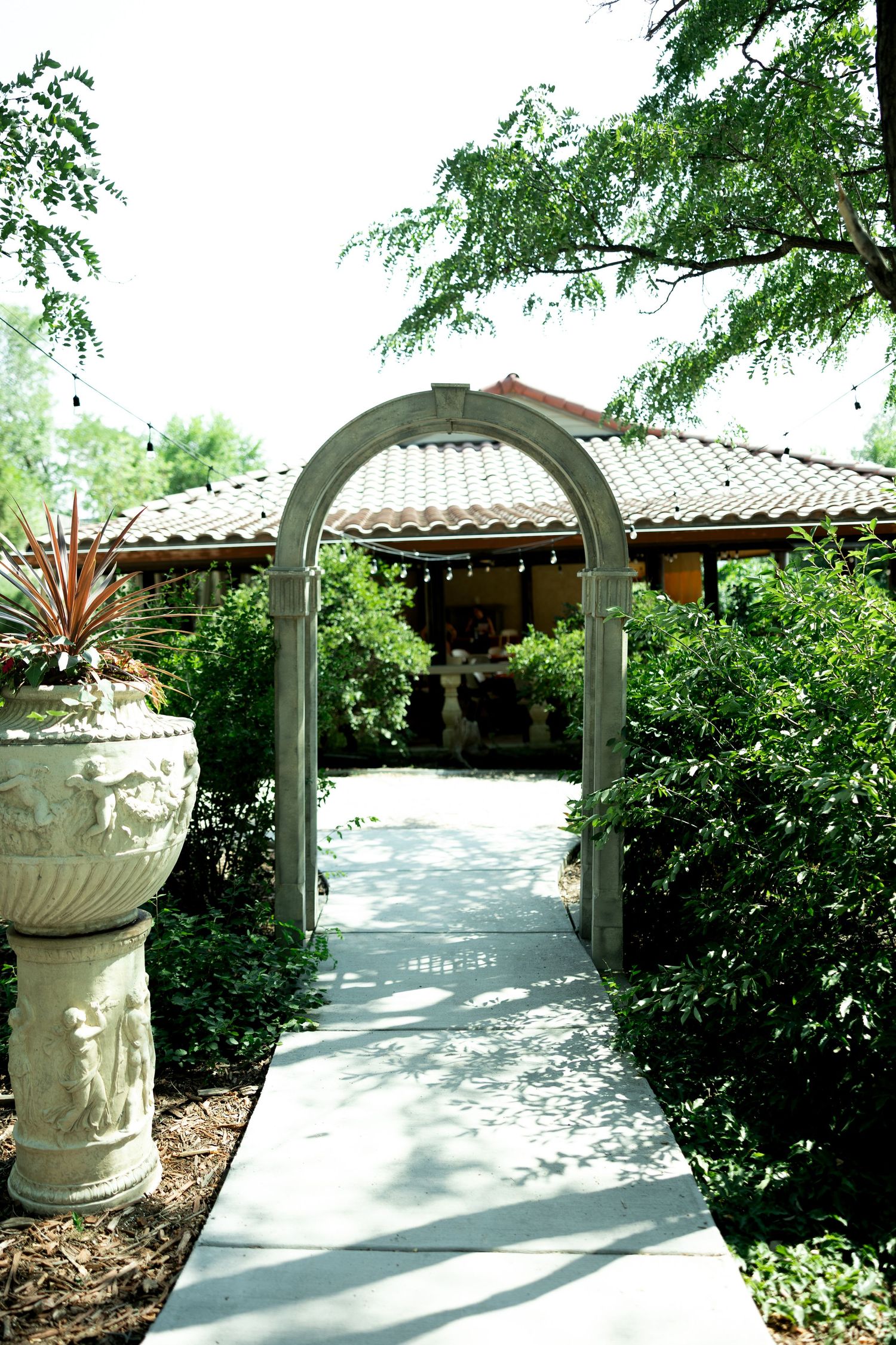 A stone archway leads down a concrete path to a Spanish-style venue surrounded by lush green hedges and trees.