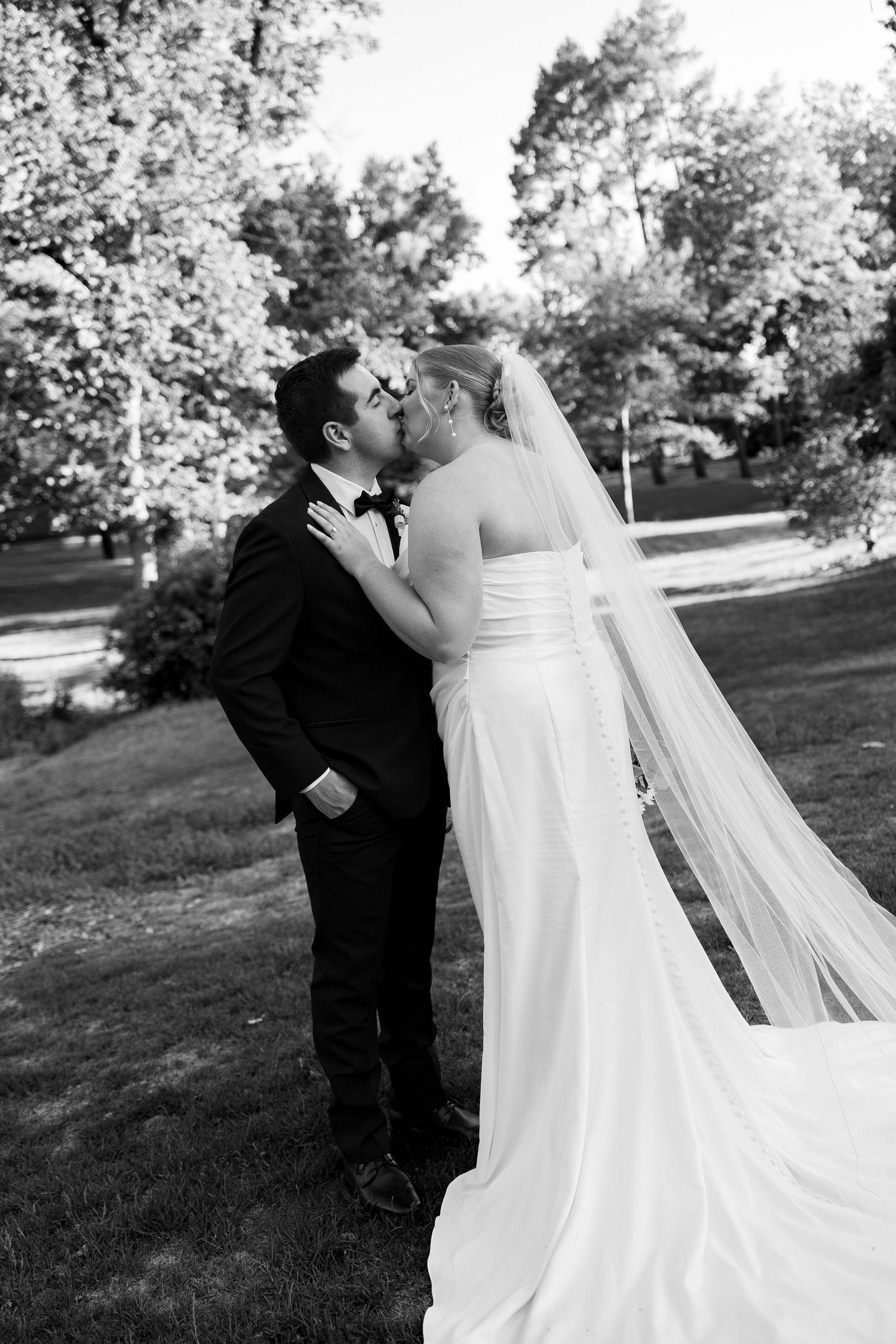 Black and white photo of a bride and groom sharing a kiss outdoors, with trees in the background.