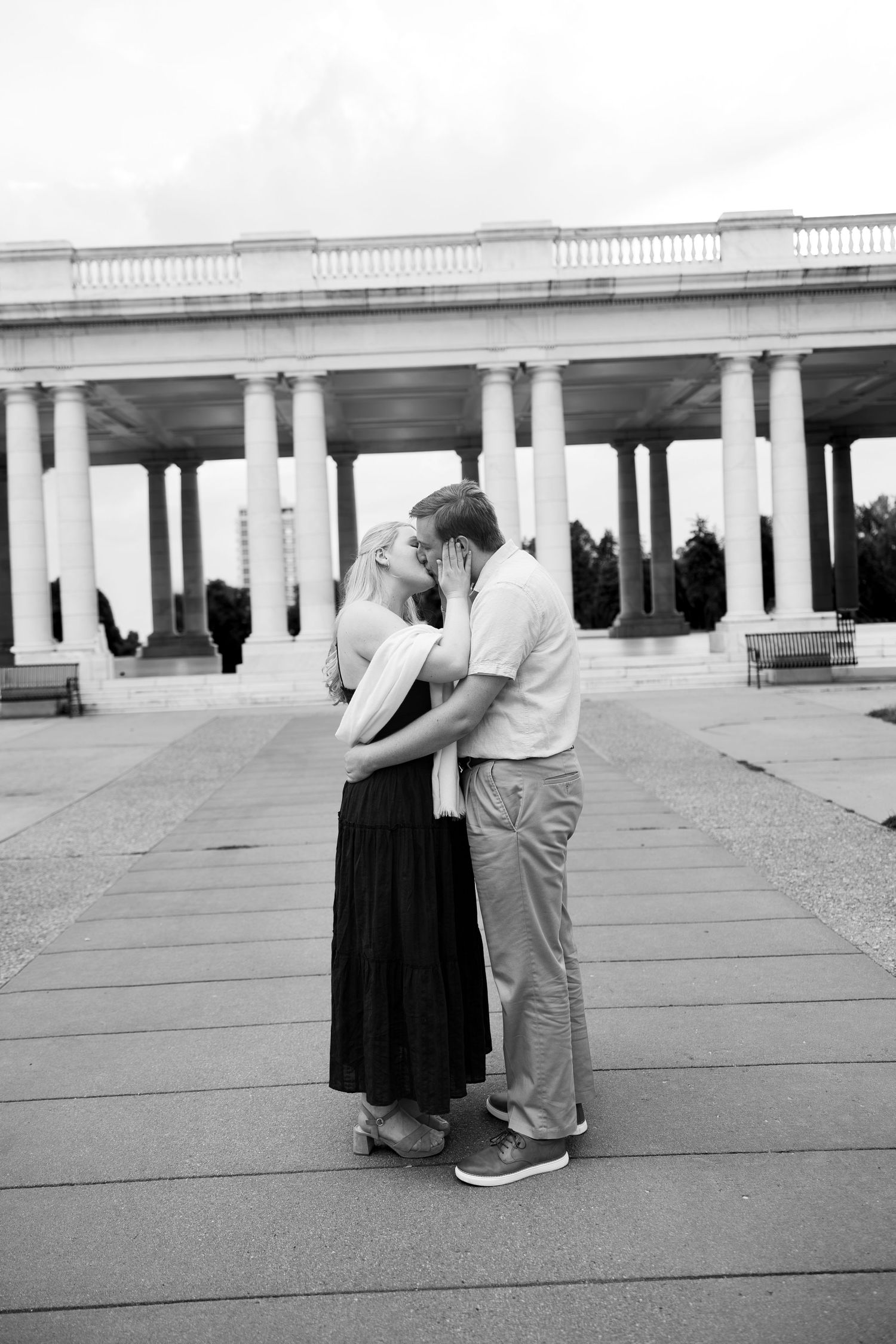 Black and white photo of a romantic couple kissing in front of a grand colonnade with white pillars.