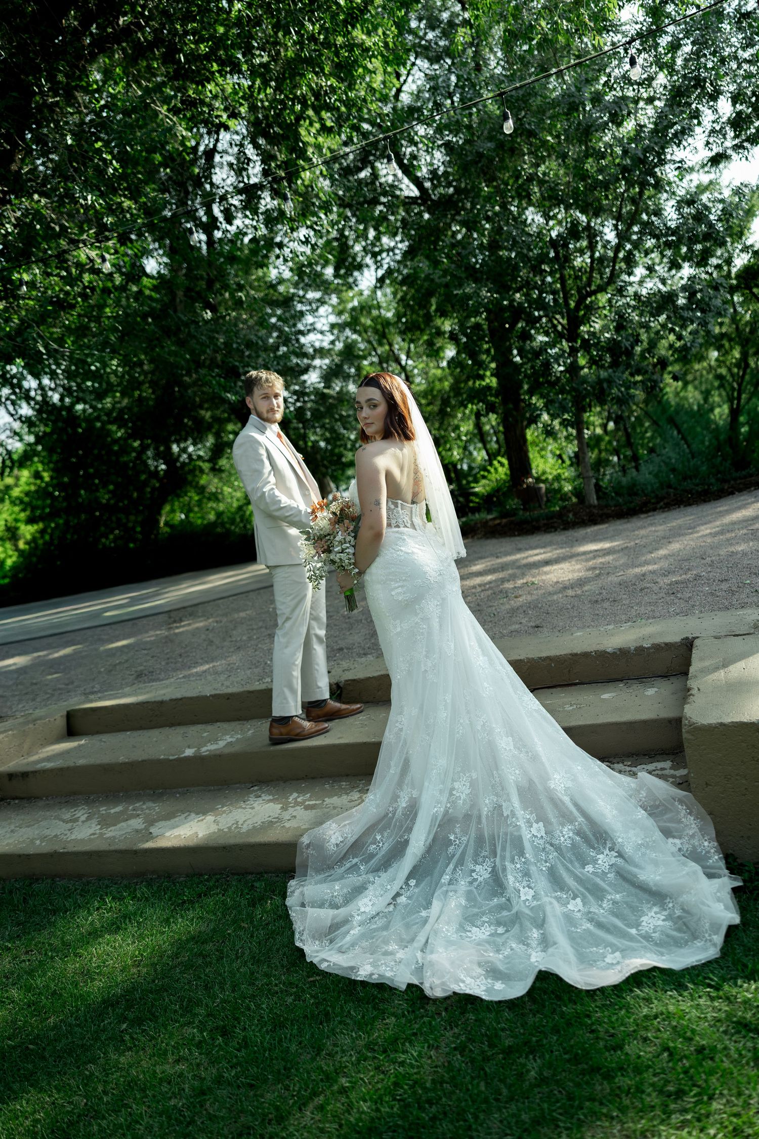 Bride in white lace gown with long train stands with groom in beige suit on outdoor steps surrounded by greenery.