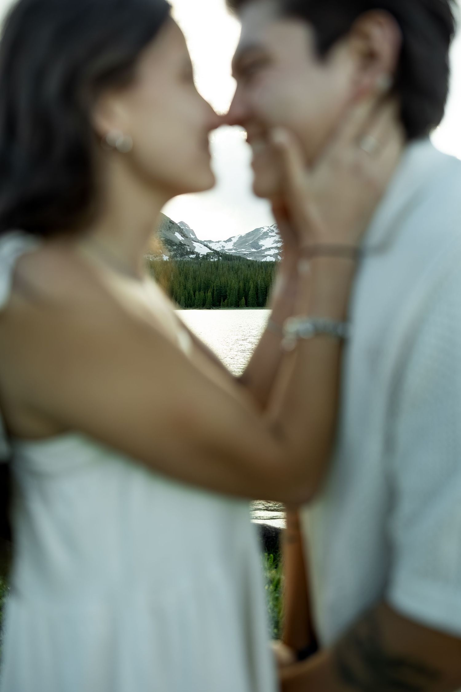 Couple sharing a romantic kiss outdoors near a mountain lake surrounded by pine trees and snow-capped peaks.