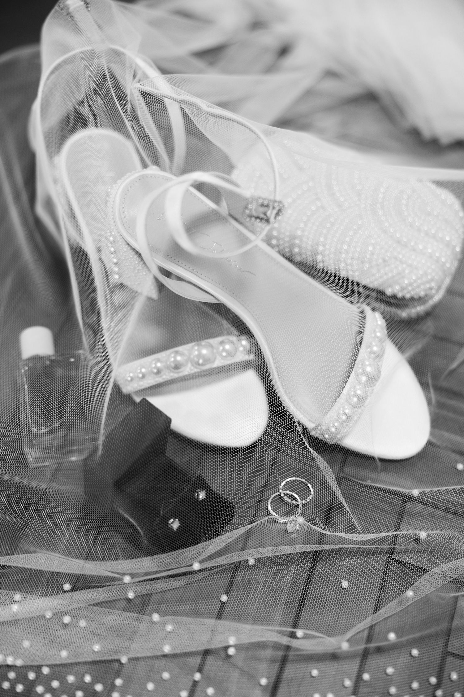 Black and white photo of bridal heels with pearl details, wedding rings, perfume, and veil arrangement.