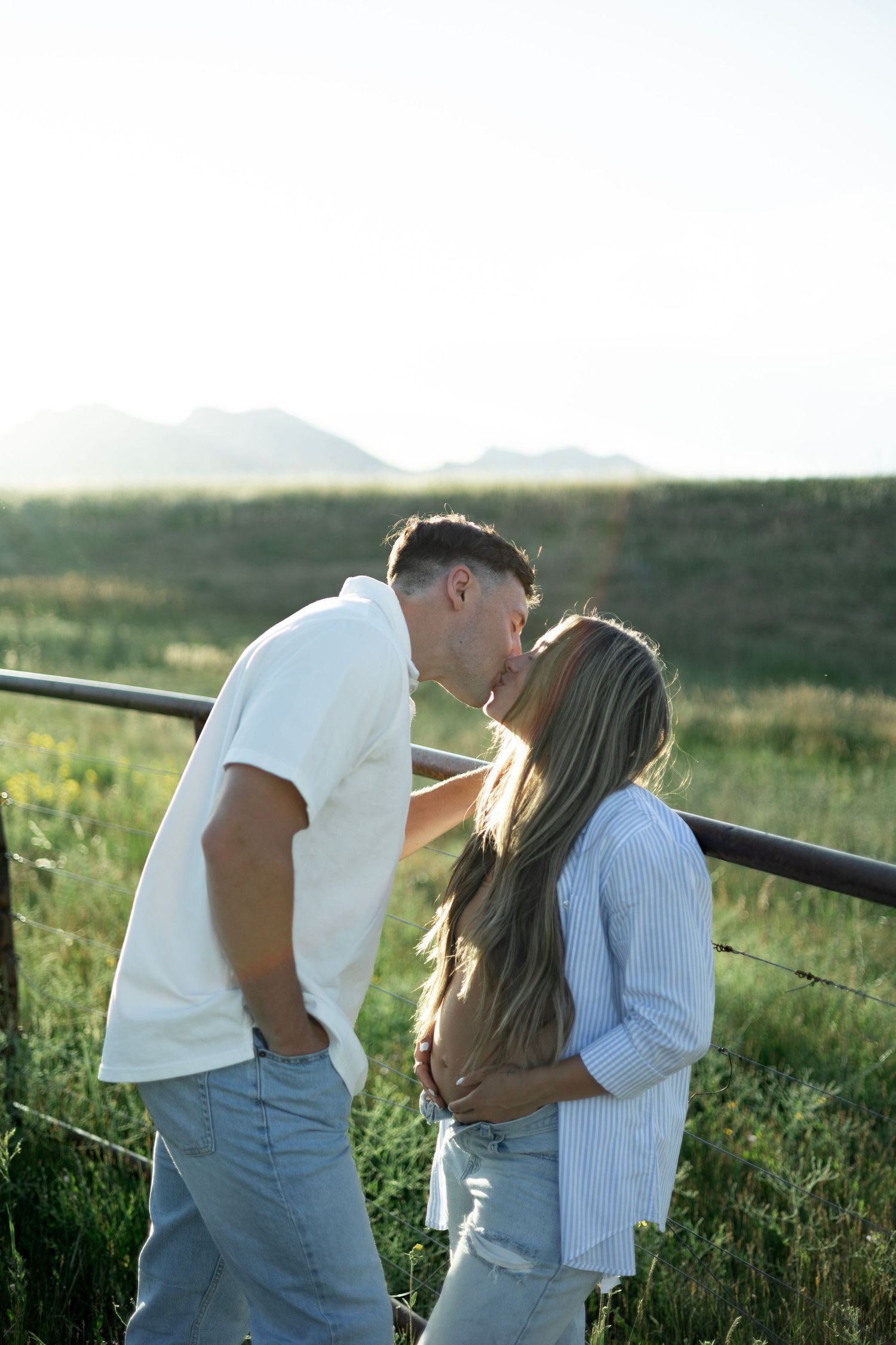 A romantic couple embraces in a field at sunset with mountains in the background.