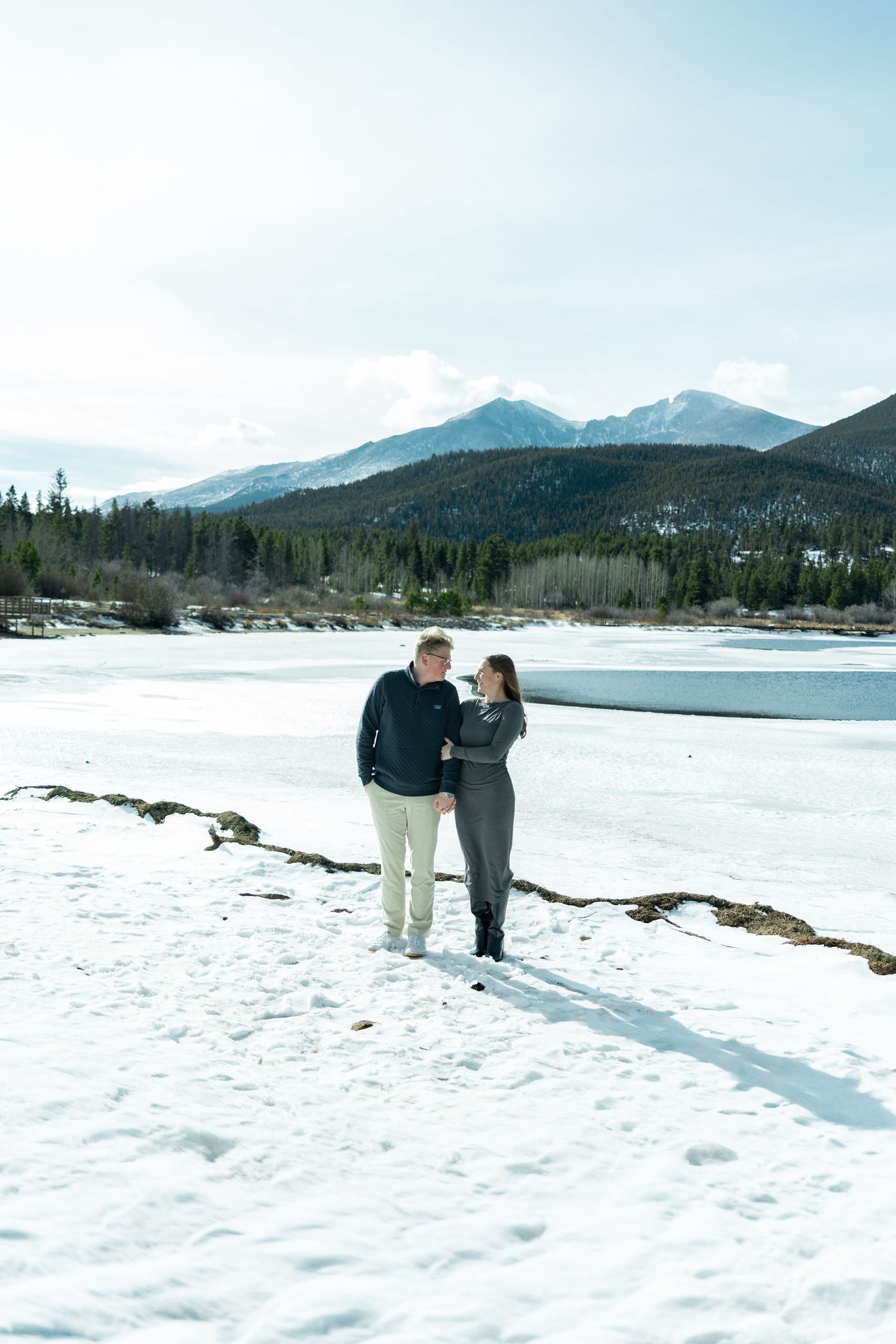 Couple standing together on snowy shore gazing at each other with mountain and frozen lake views.
