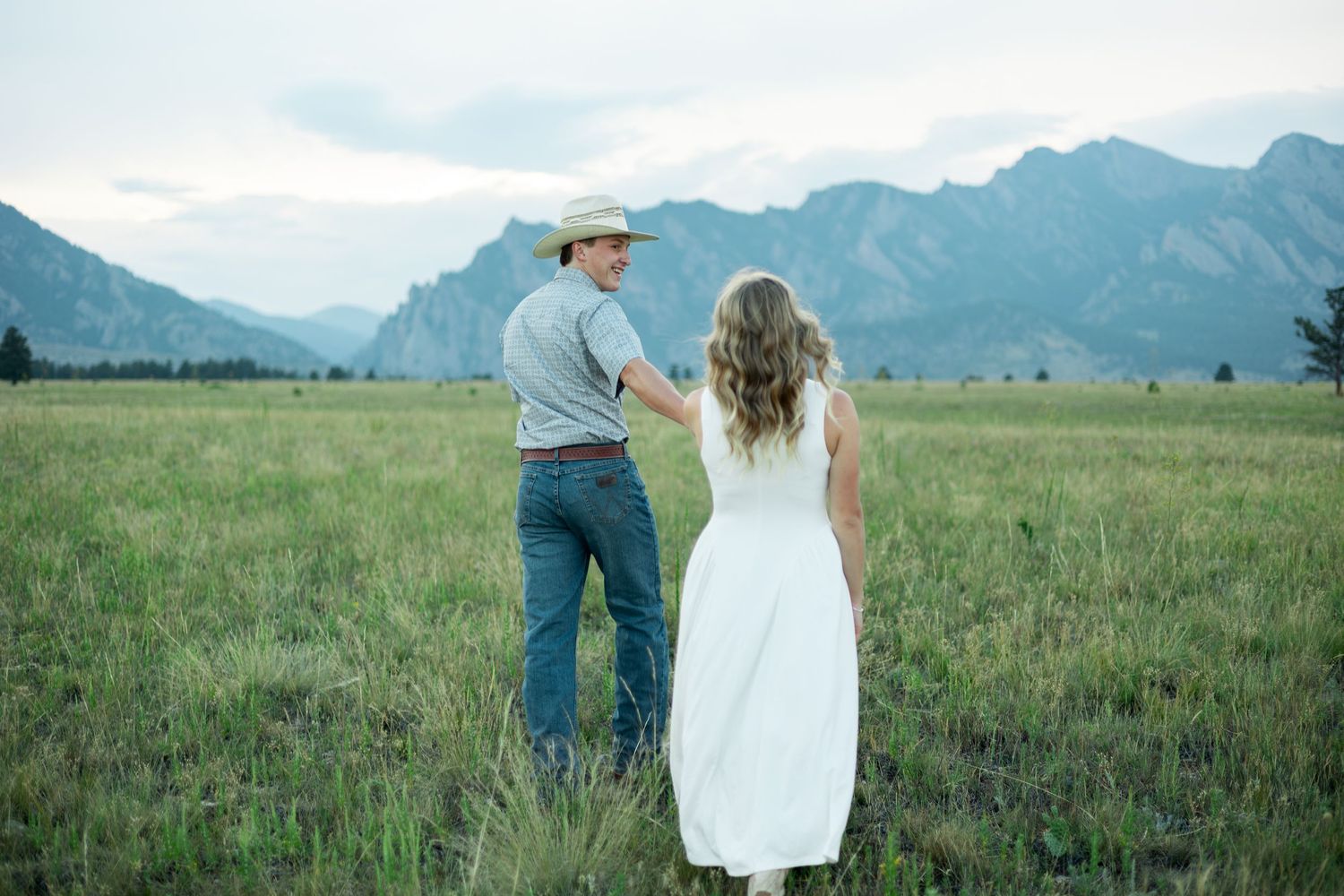 Couple holding hands in a green meadow with stunning Rocky Mountain backdrop, man in cowboy hat and woman in white dress.