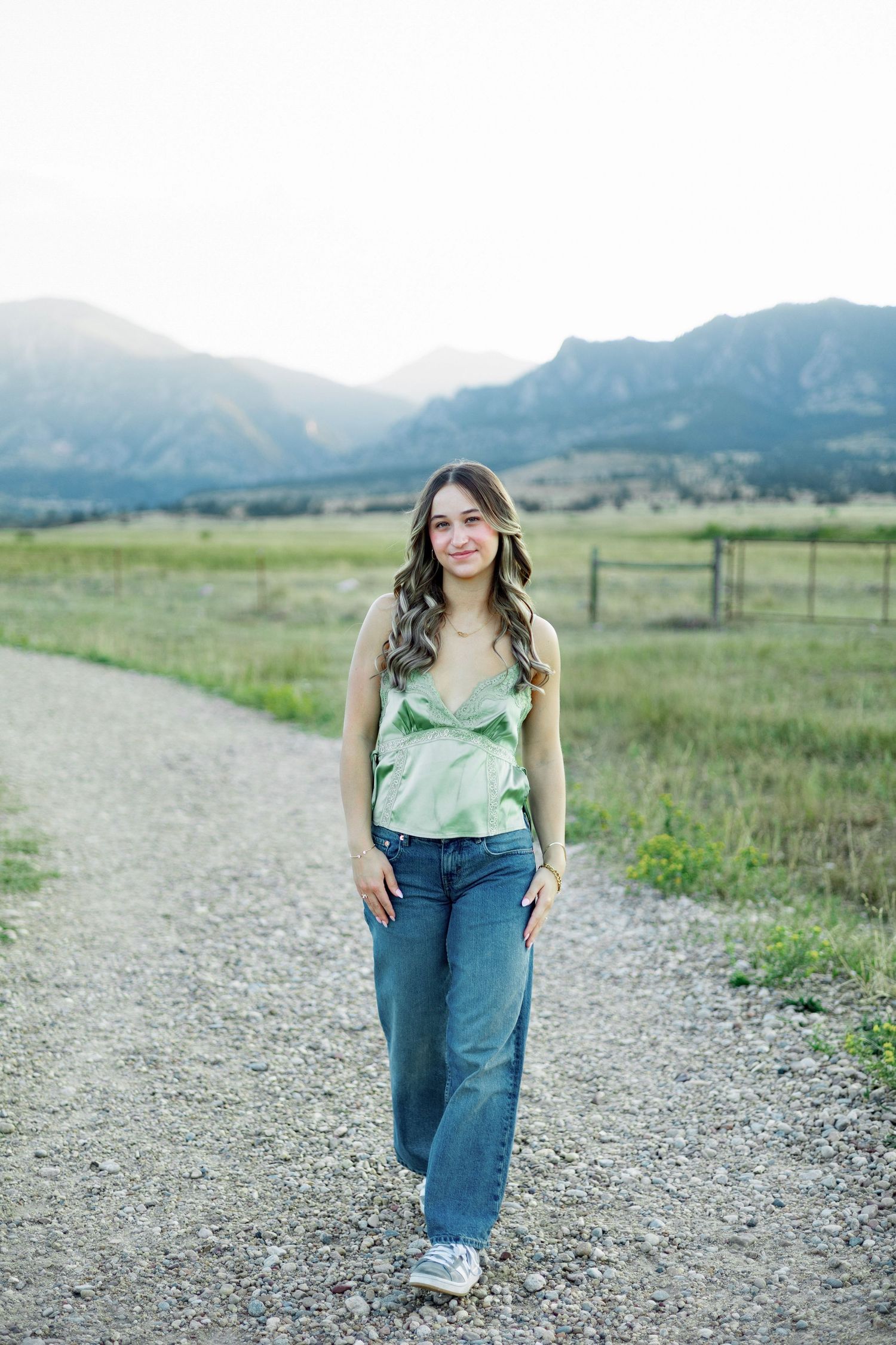 Young woman in green top and jeans walks a gravel path with scenic mountain backdrop in Colorado.