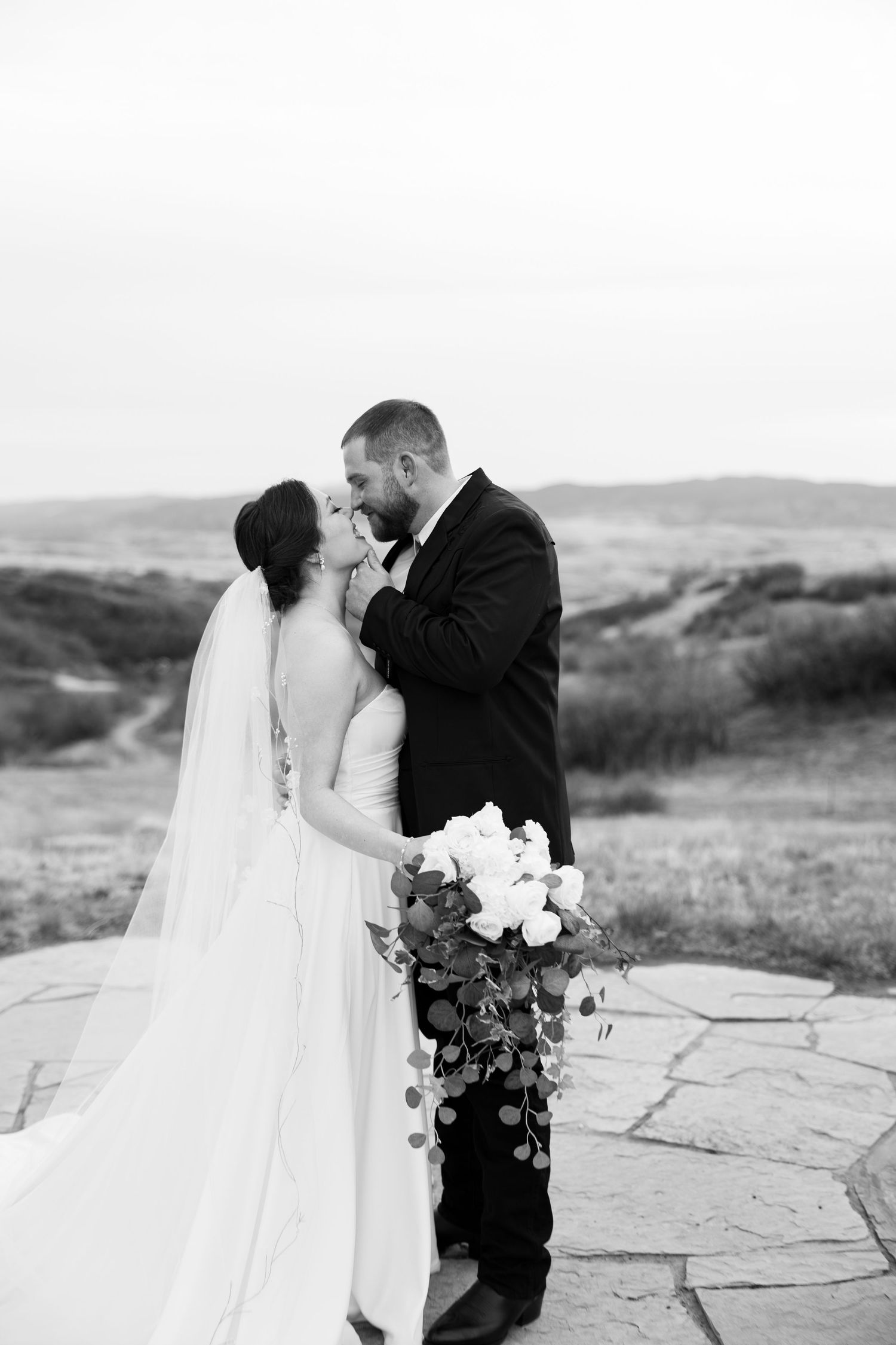 Black and white portrait of bride and groom nearly kissing on stone patio with scenic countryside backdrop.